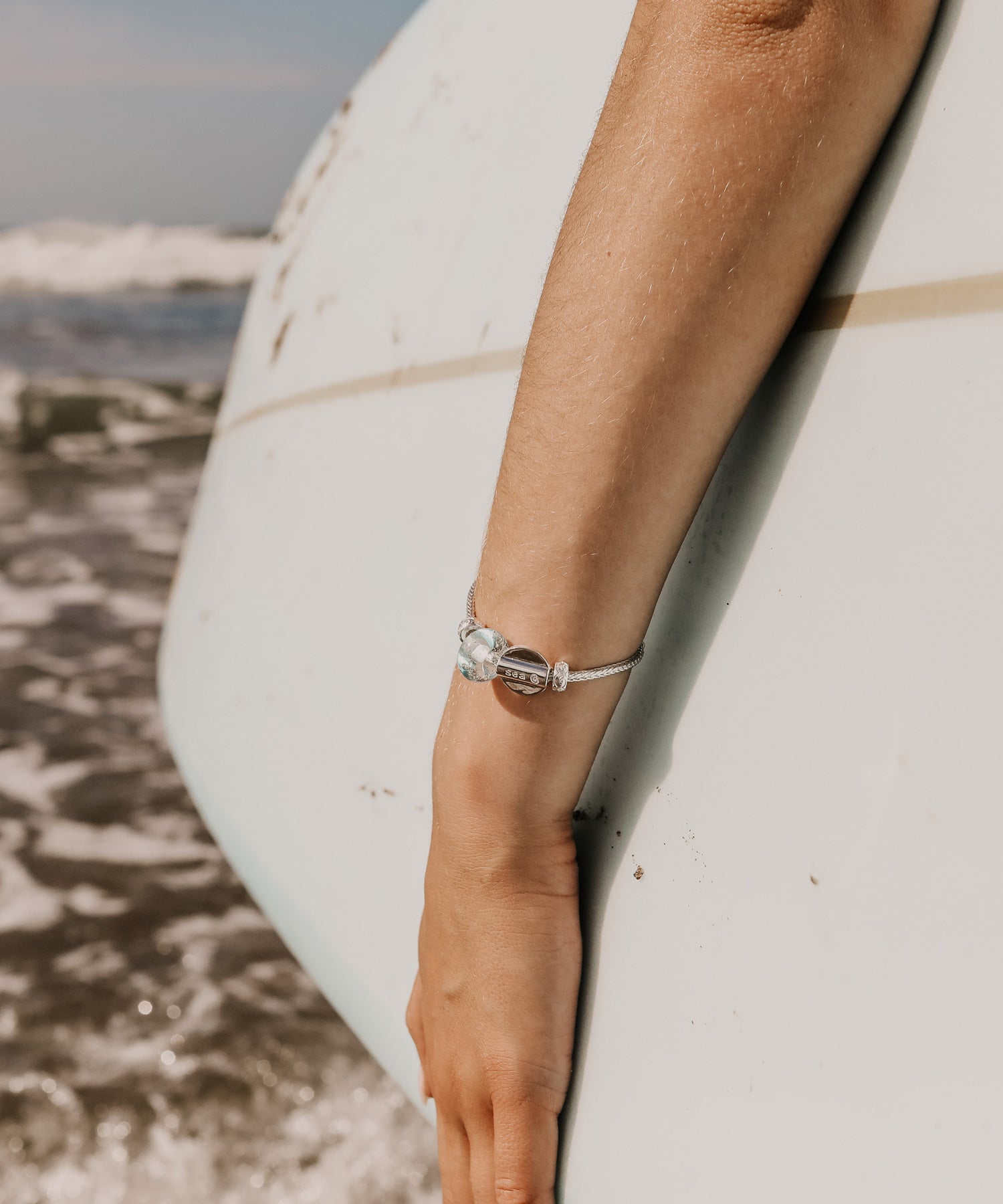 Woman carrying surfboard wearing silver bracelet with baby blue beach sand bead on Croyde Bay, North Devon.