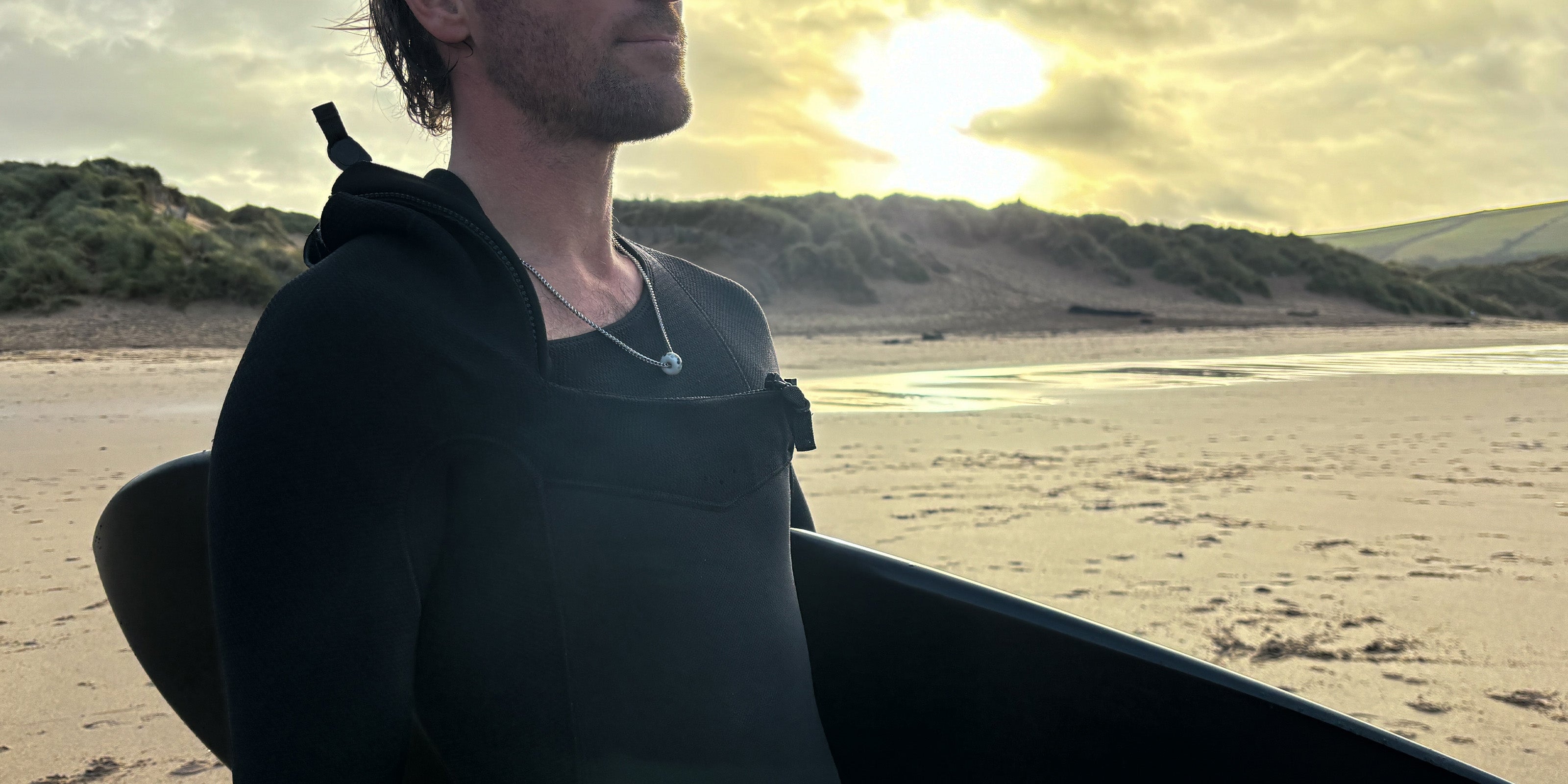 Man with surfboard on the beach at sunrise wearing a Nalu chain bead necklace.