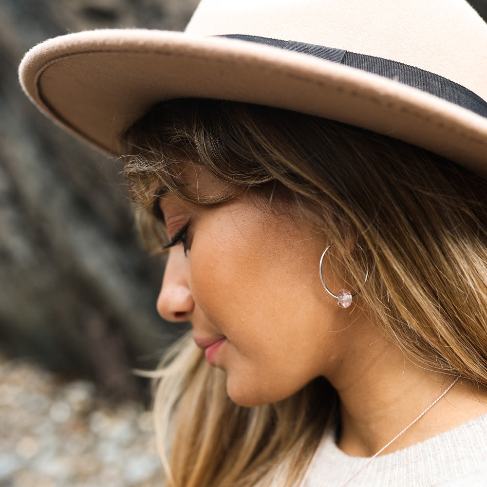 Woman wearing a hat and large silver hoop earrings with pale pink beach drop glass bead on Croyde Bay beach.