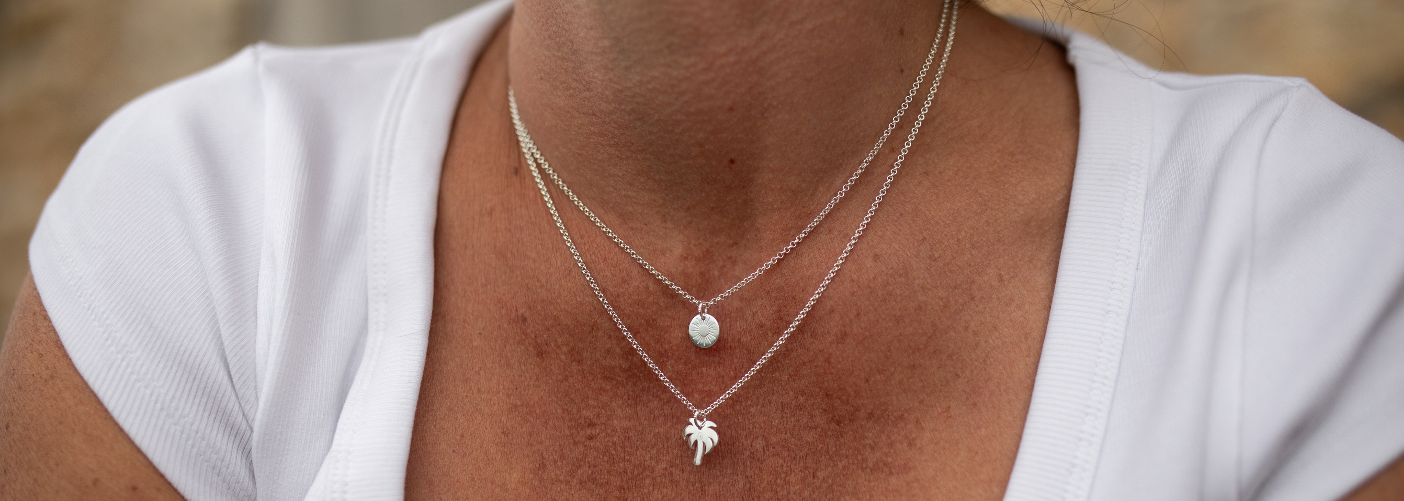 A close-up of a person wearing a double-layered silver necklace. The top layer features a delicate chain with a small round disc pendant, and the bottom layer has a slightly thicker chain with a small palm tree pendant.
