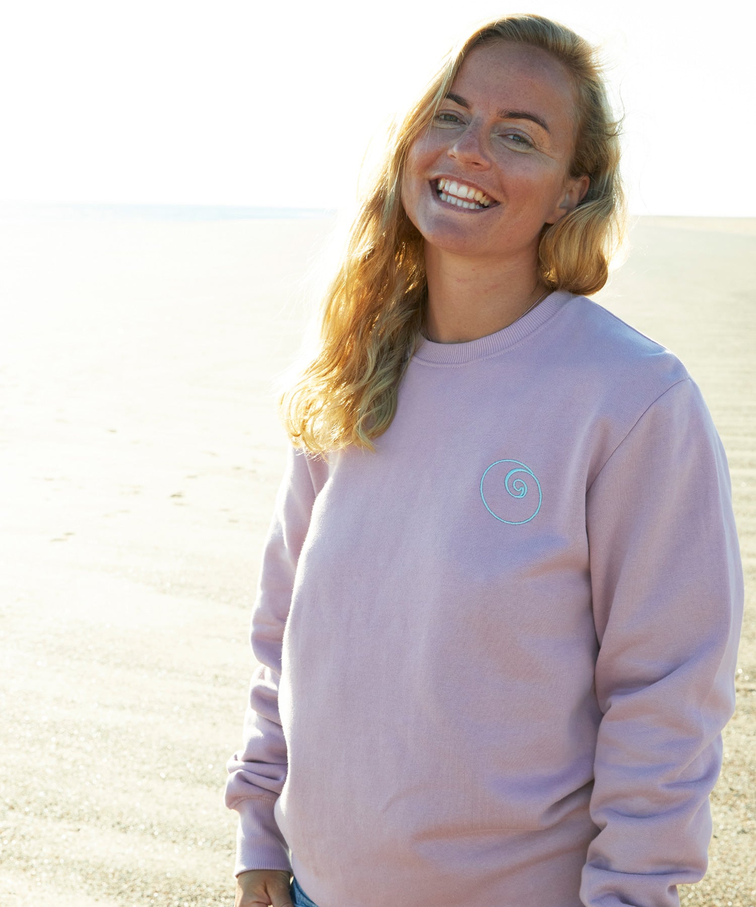 Woman standing on the beach in a dusty pink blush organic cotton Nalu sweatshirt.