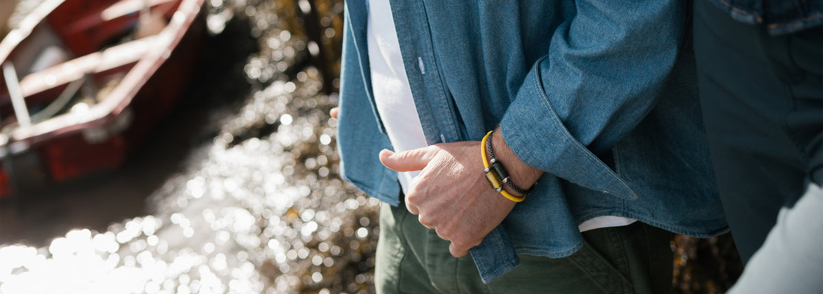 A person outdoors wearing a blue denim button-up shirt over a white T-shirt, with olive green pants and a yellow bracelet, standing near water and a red boat.