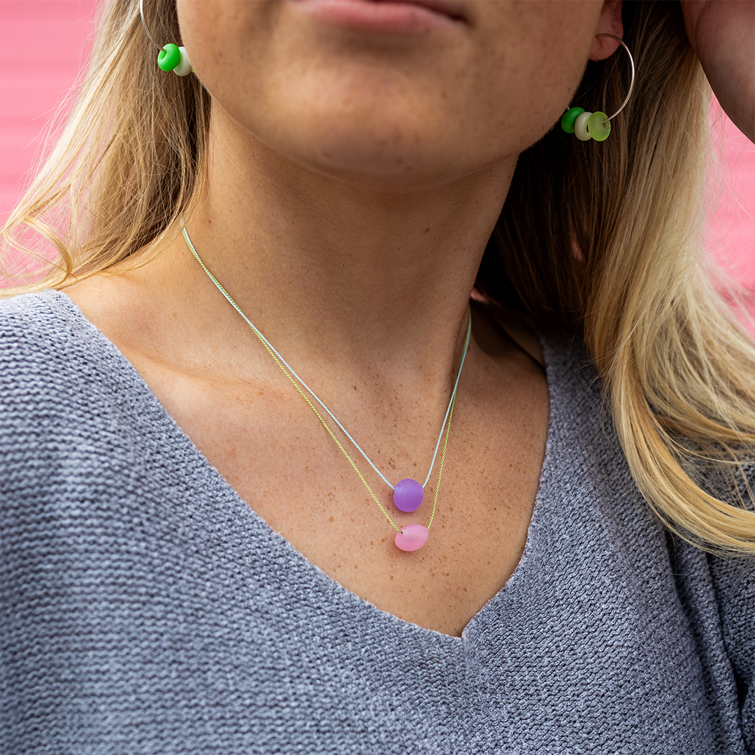 Blonde haired woman in grey sweater wearing silk necklaces in pink and purple.