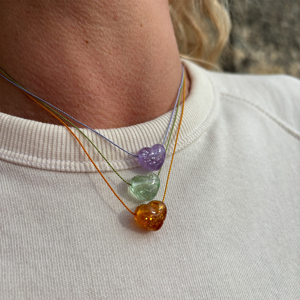 Purple, green and amber heart glass pendant necklaces on person wearing a white shirt.