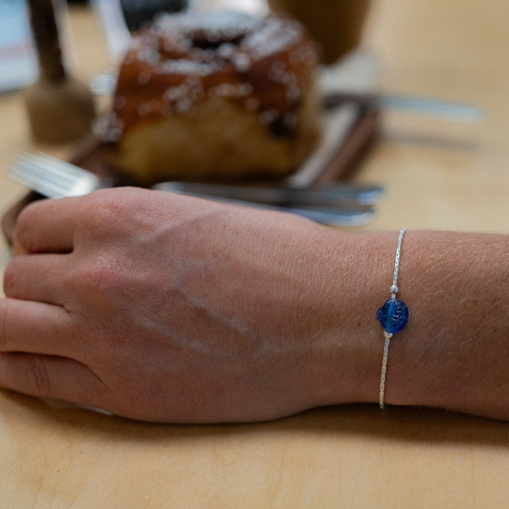 Hand wearing a blue and silver bracelet on a wooden surface with a blurred pastry in the background