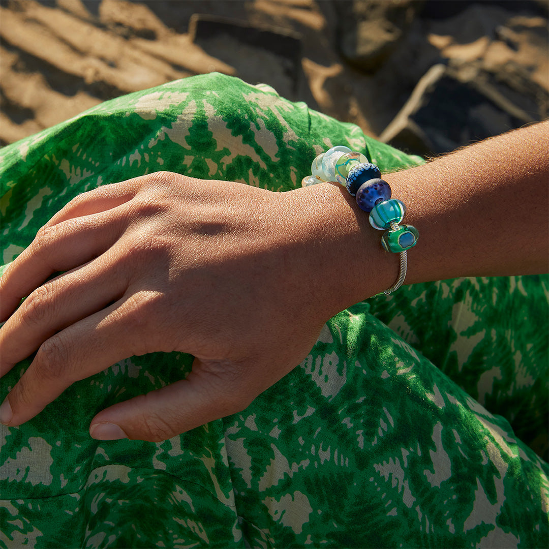 Purple, blue, green patterned glass beads representing beaches on a silver bracelet worn by woman in green skirt.