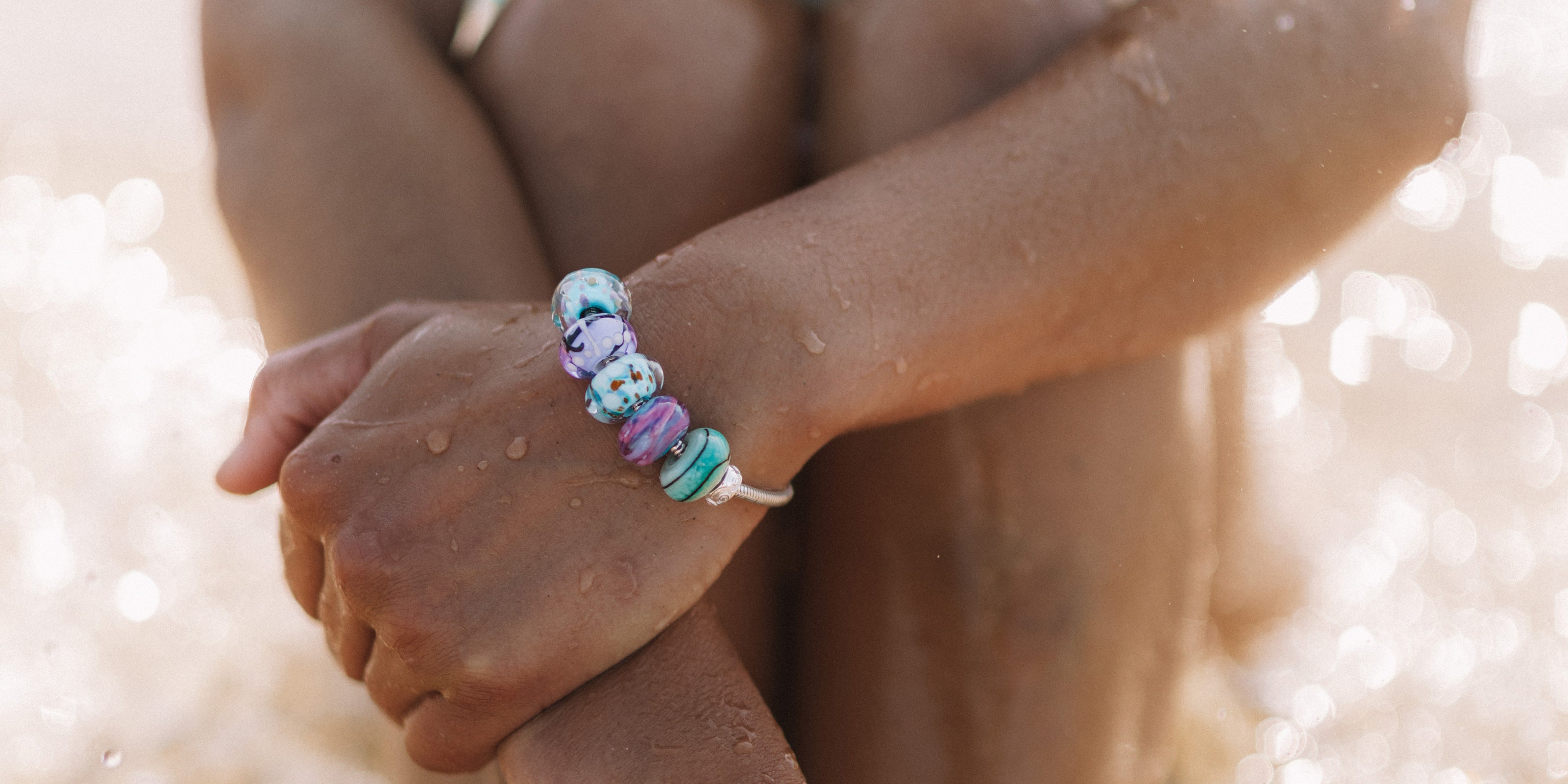 Green, blue and purple glass beads worn on silver snake chain bracelet by surfer girl at beach.