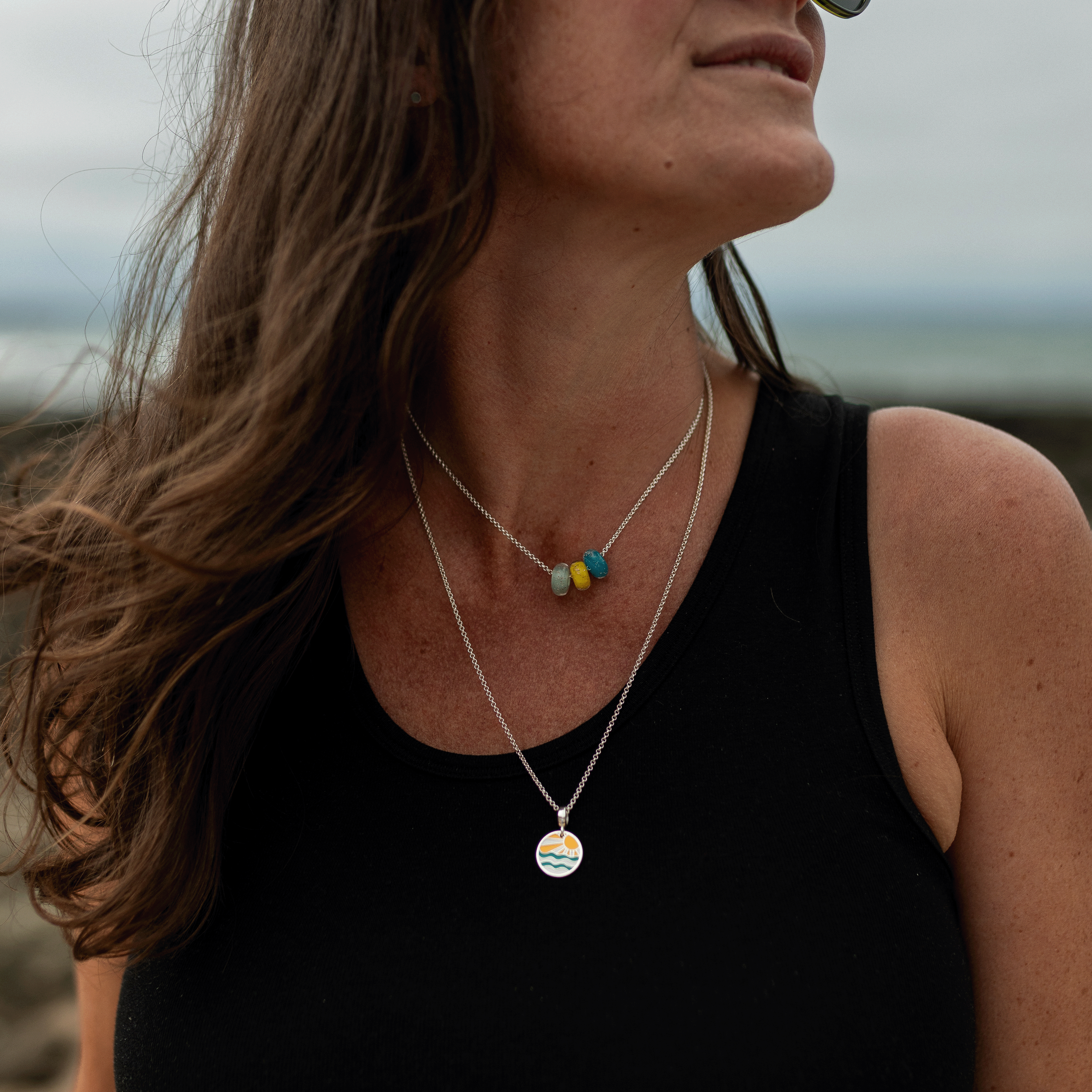 A woman wearing layered silver necklaces, one with three small colorful beads and the other with a round pendant, photographed outdoors with a blurred natural background.