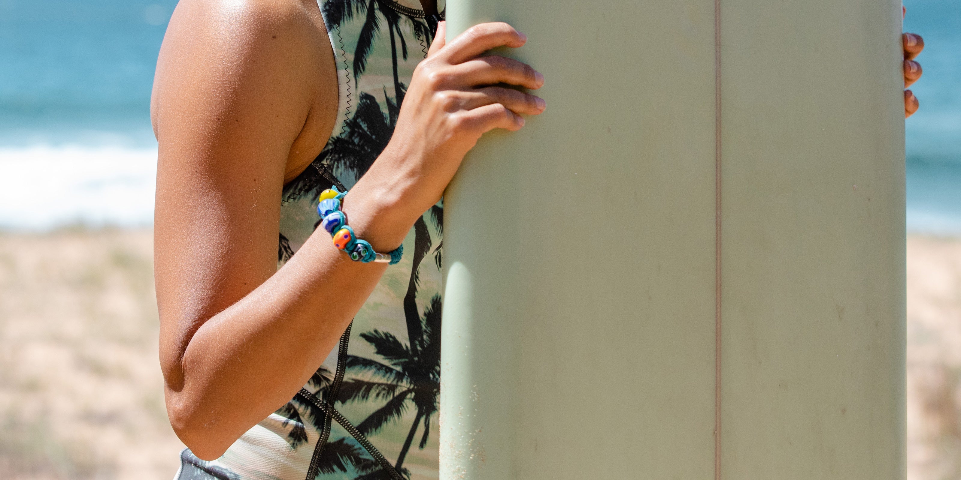 Custom macrame bracelet with colourful glass beads in teal cord, worn by surfer on the beach.