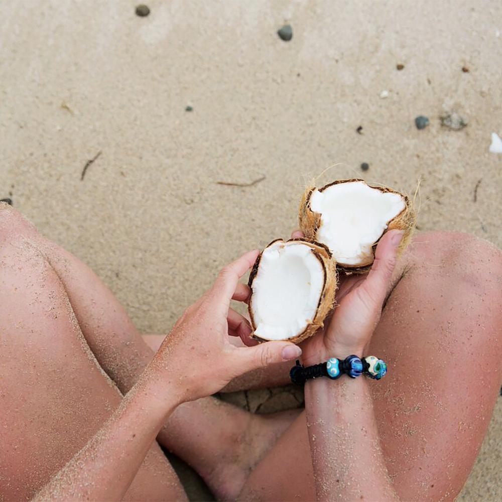 Person sitting on the beach holding coconut, wearing a black cord bead bracelet.