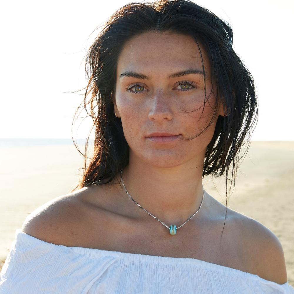 Woman with long dark hair, on the beach wearing a white top and glass bead on silver chain necklace