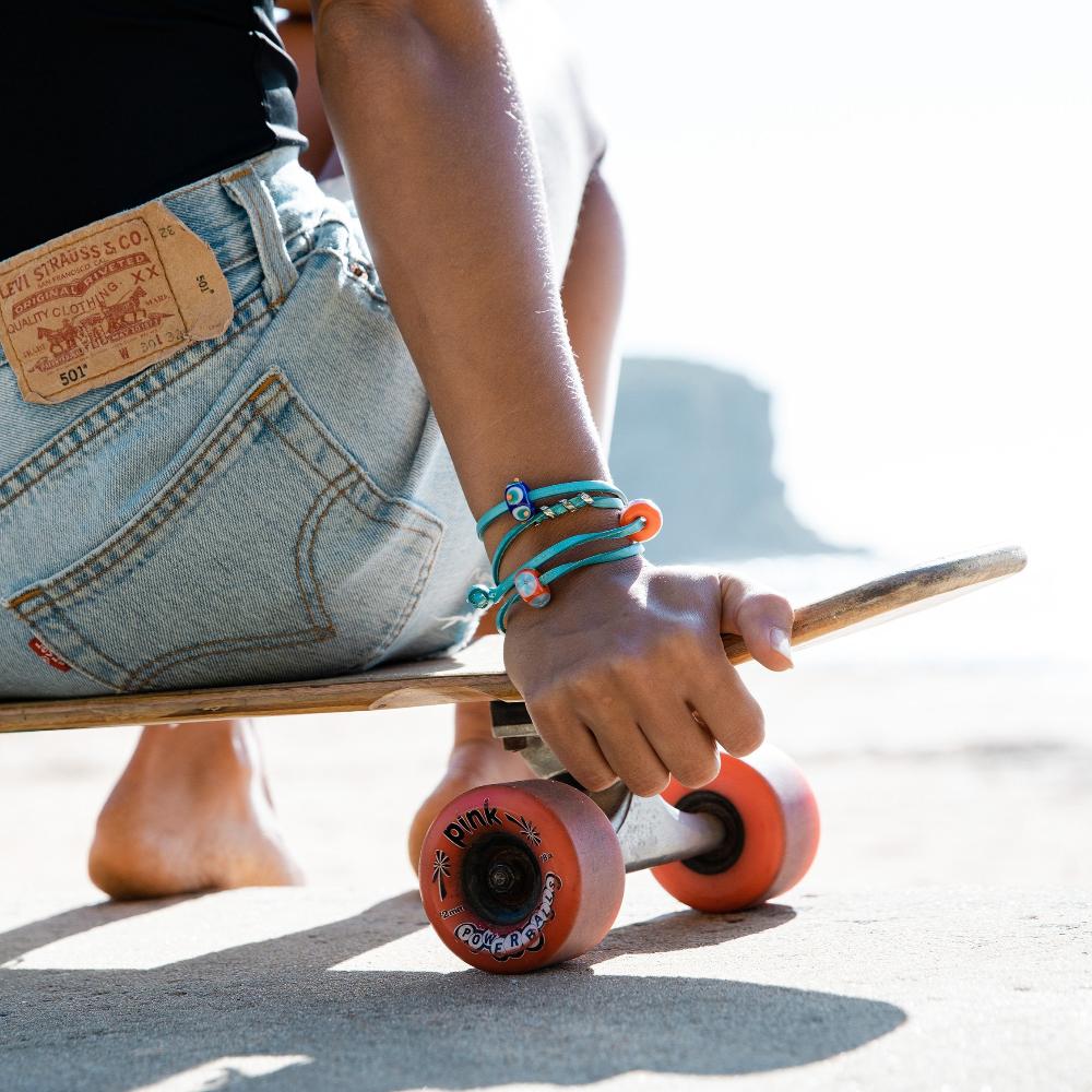 Woman wearing denim shorts, sitting on a skateboard with orange wheels, wearing a turquoise leather bracelet with silver and orange glass beads.