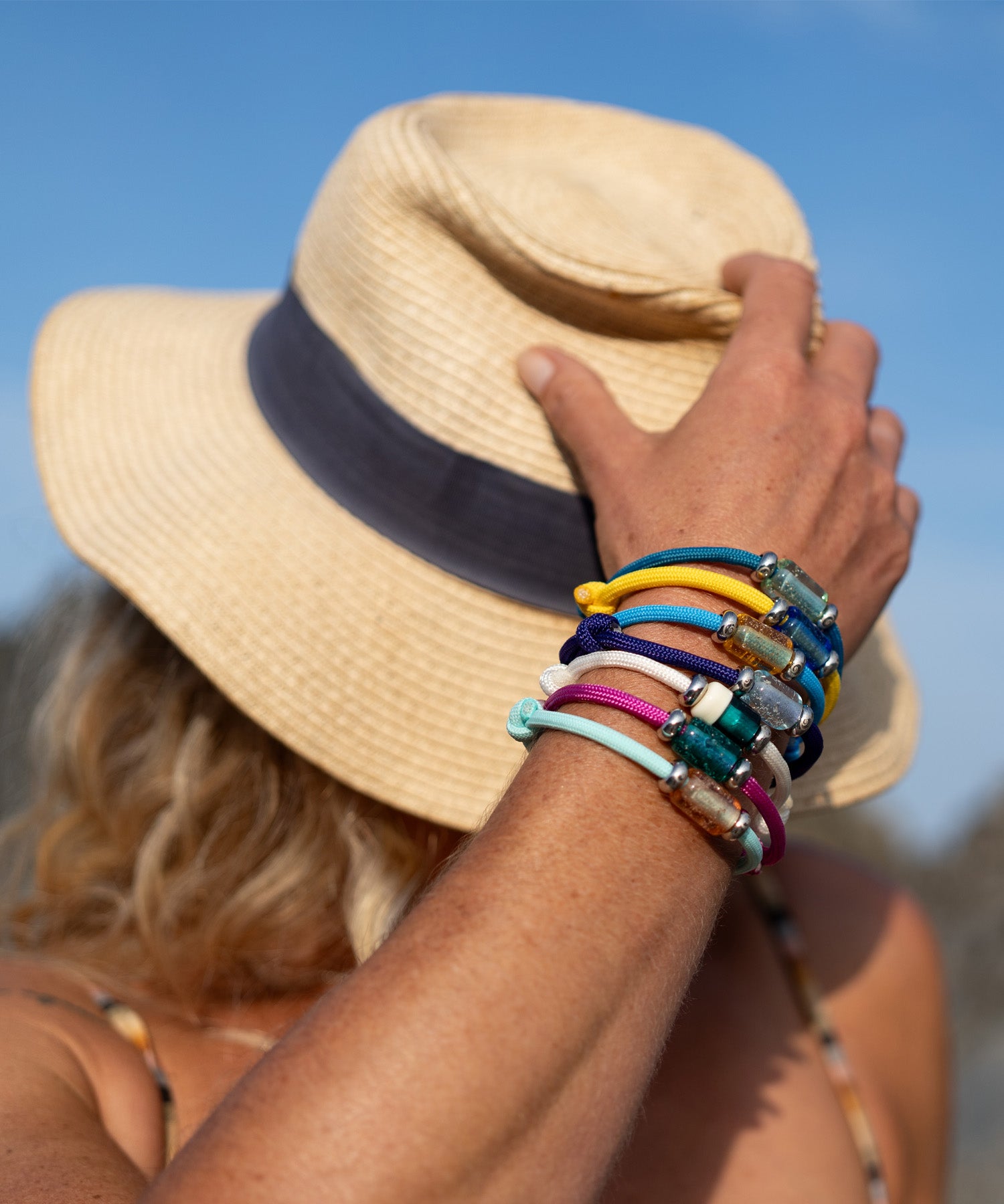 Person wearing a straw hat with a blue band, holding it with a hand adorned with colorful bracelets against a clear blue sky.