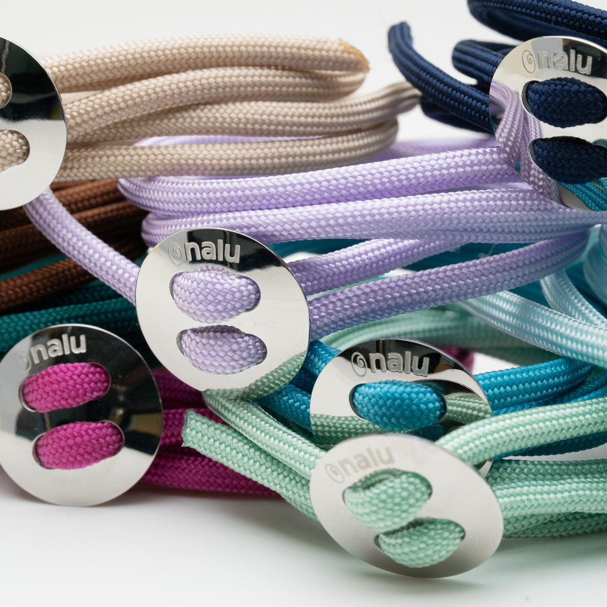 A close-up pile of braided bracelets with shiny oval metal clasps branded 'nalu'. The ties are shown in multiple pastel and bright colors including beige, navy, lavender, teal, mint, pink, and brown, arranged on a clean white background.