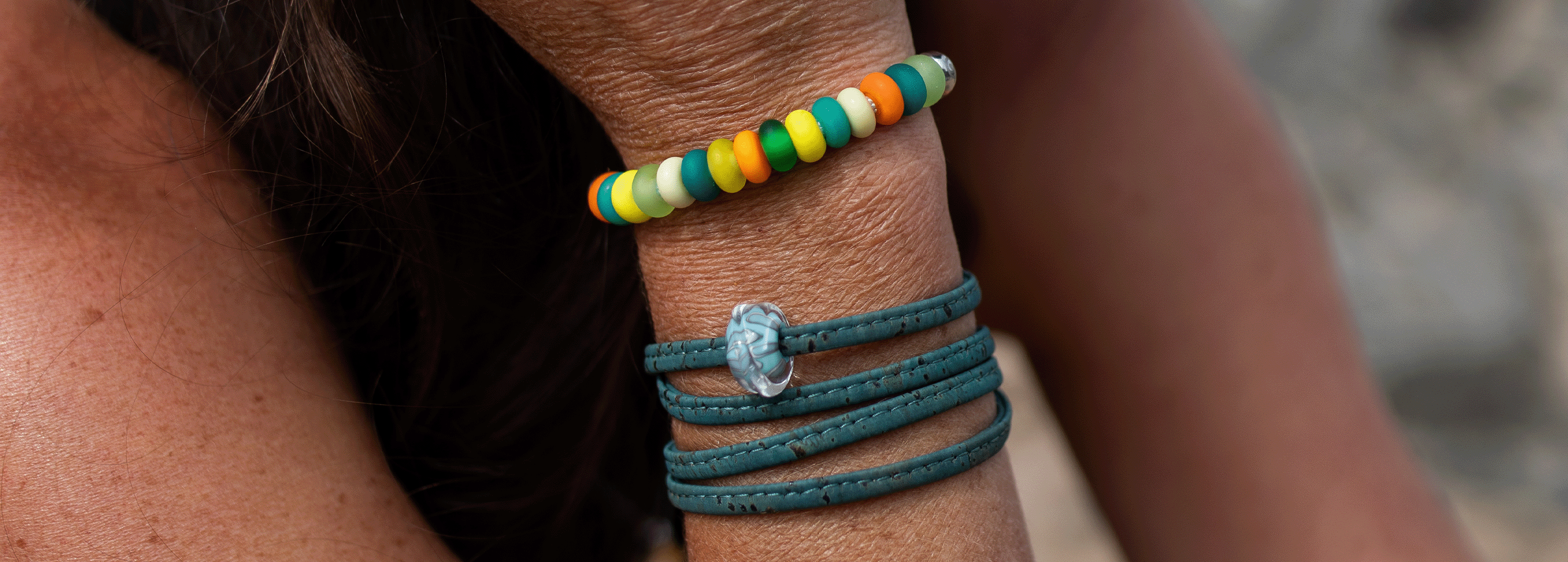 A close-up of a person's wrist wearing two bracelets: a colorful beaded bracelet with green, yellow, orange, and white beads, and a dark teal wrap bracelet with a small clear shell charm.