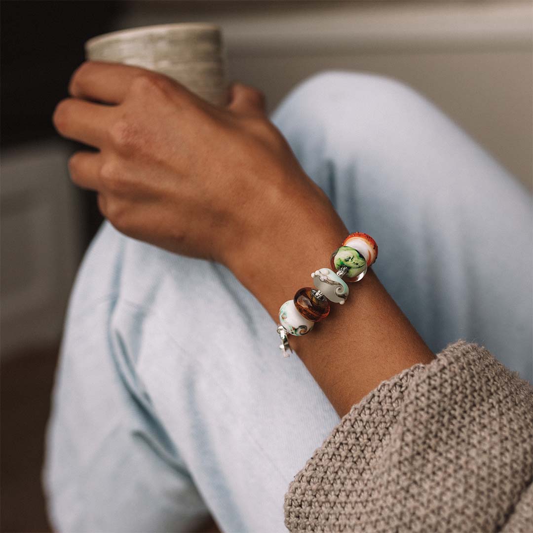 Person wearing jeans and knitted jumper, holding a cup of coffee, wearing a silver bracelet with colourful glass beads.