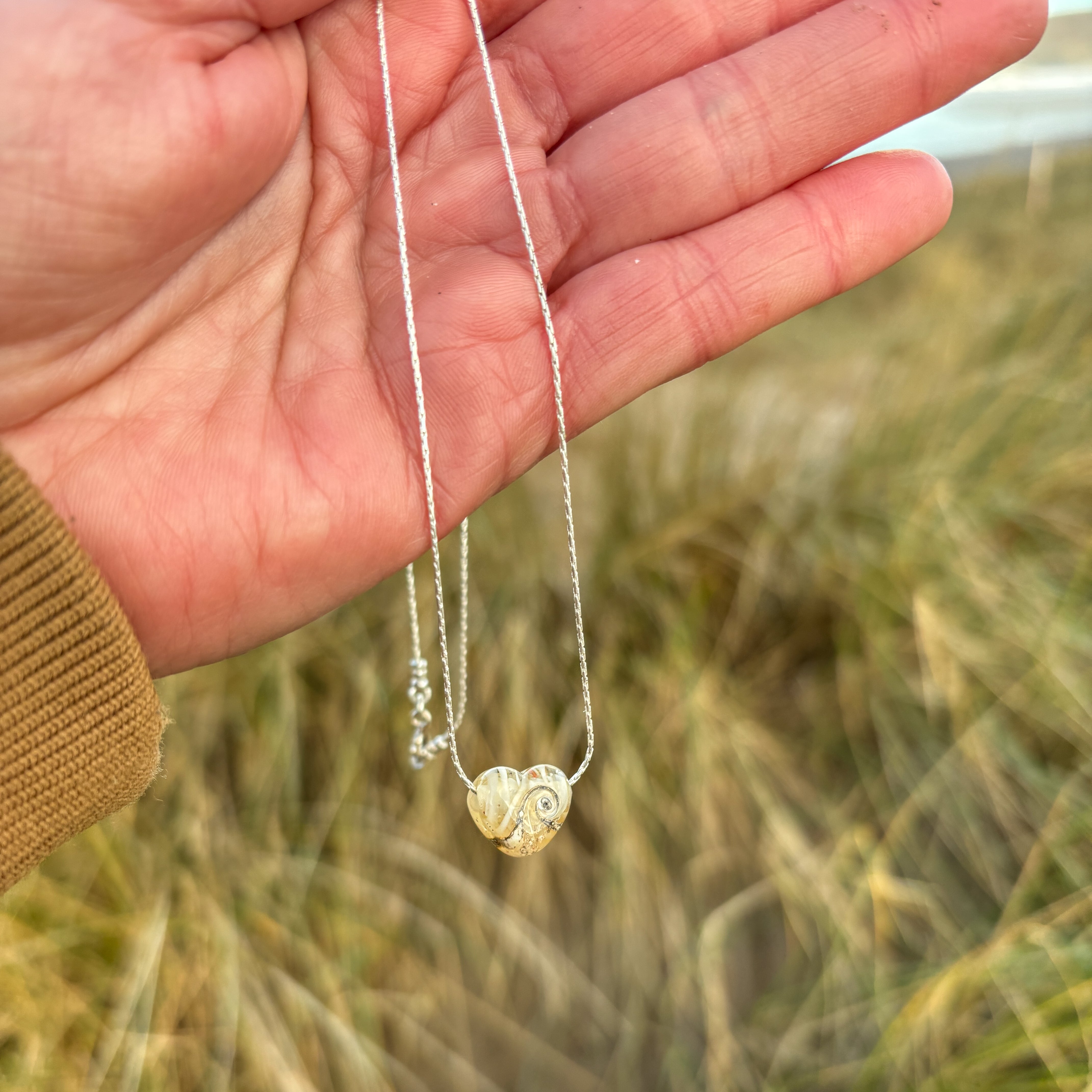 Cream sand heart on silver chain held in persons hand at the beach.