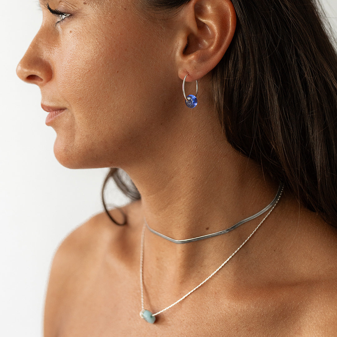 Woman with dark hair wearing silver necklaces and blue beaded earring hops..