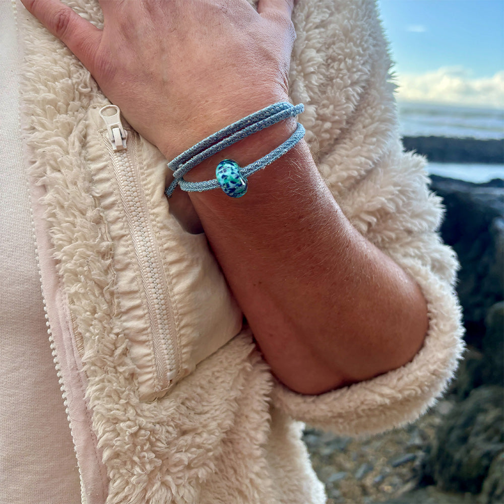 Person wearing a white fleece jacket and denim wrap bracelet with blue glass bead on it.