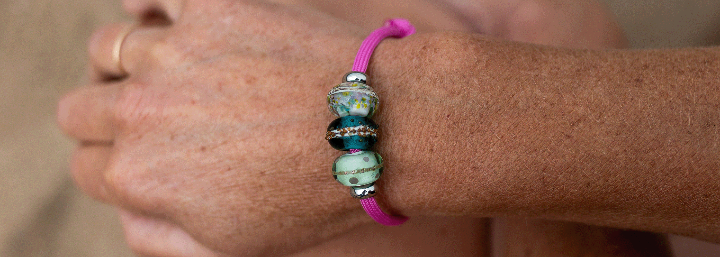 A close-up of a person's wrist wearing a handmade magenta cord bracelet with three decorative glass beads featuring blue, green, and white patterns.