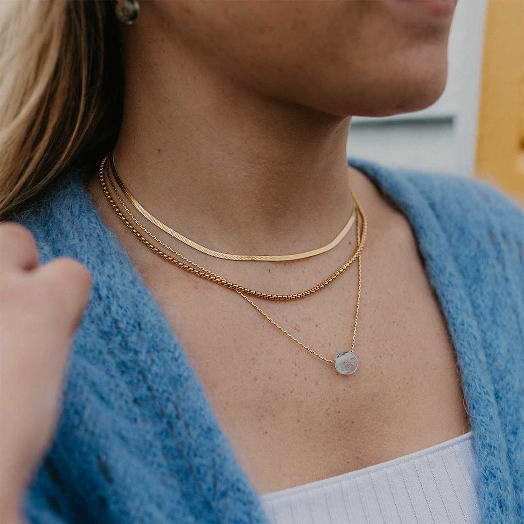 Pale blue sand pebble and gold necklace layers worn by woman in blue jumper.