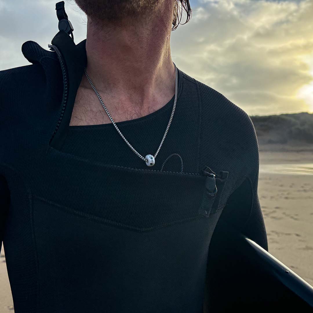 Surfer on the beach in a wetsuit wearing a silver necklace with bead.