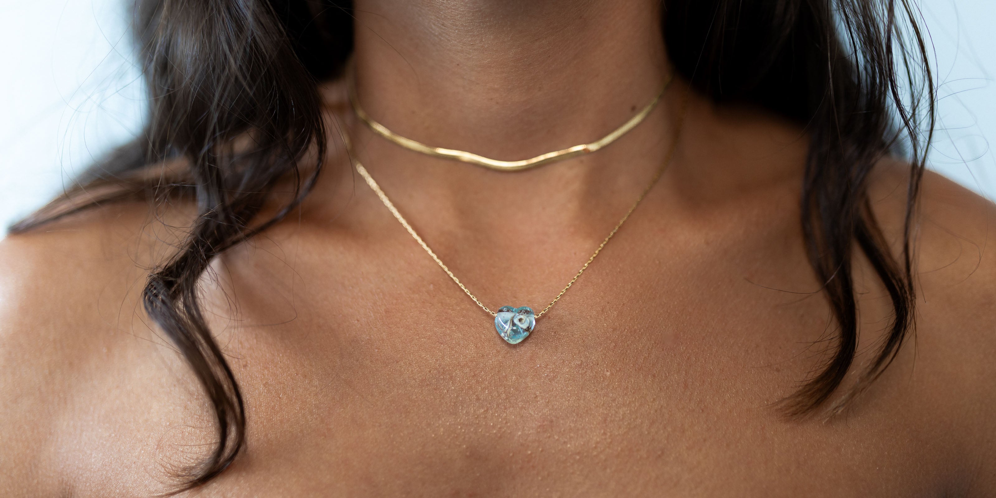 Blue mint glass heart pendant on gold chain worn by dark haired woman.