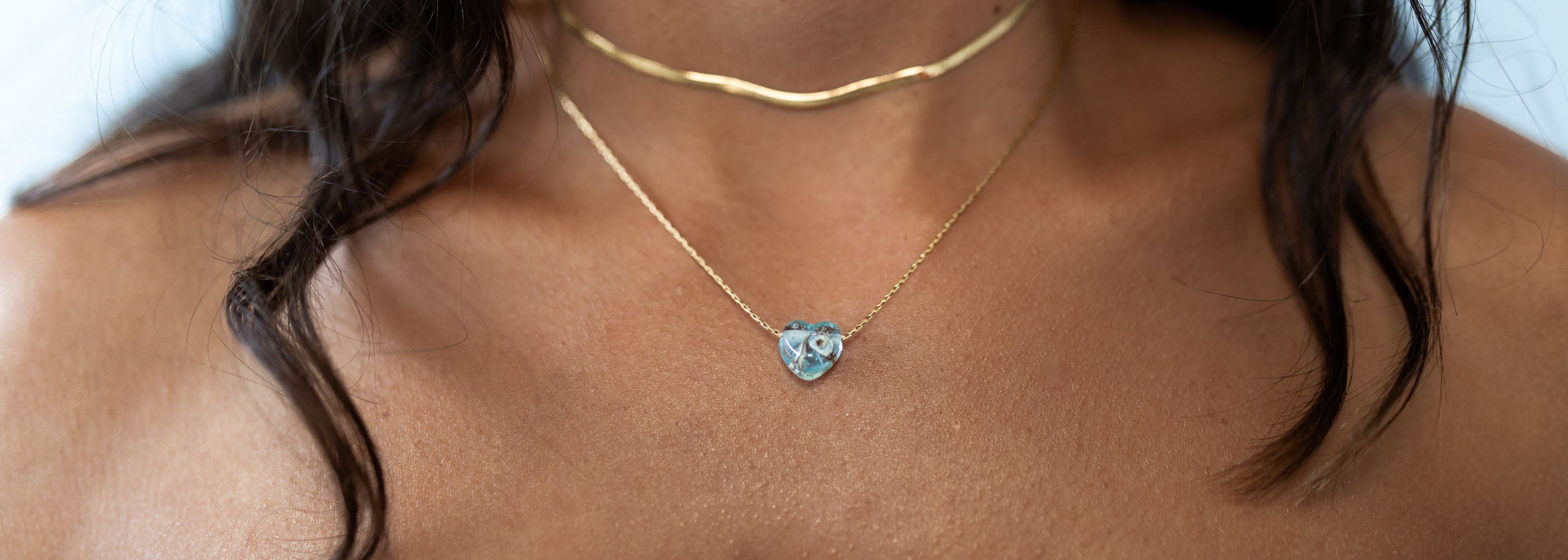 Blue mint glass heart pendant on gold chain worn by dark haired woman.