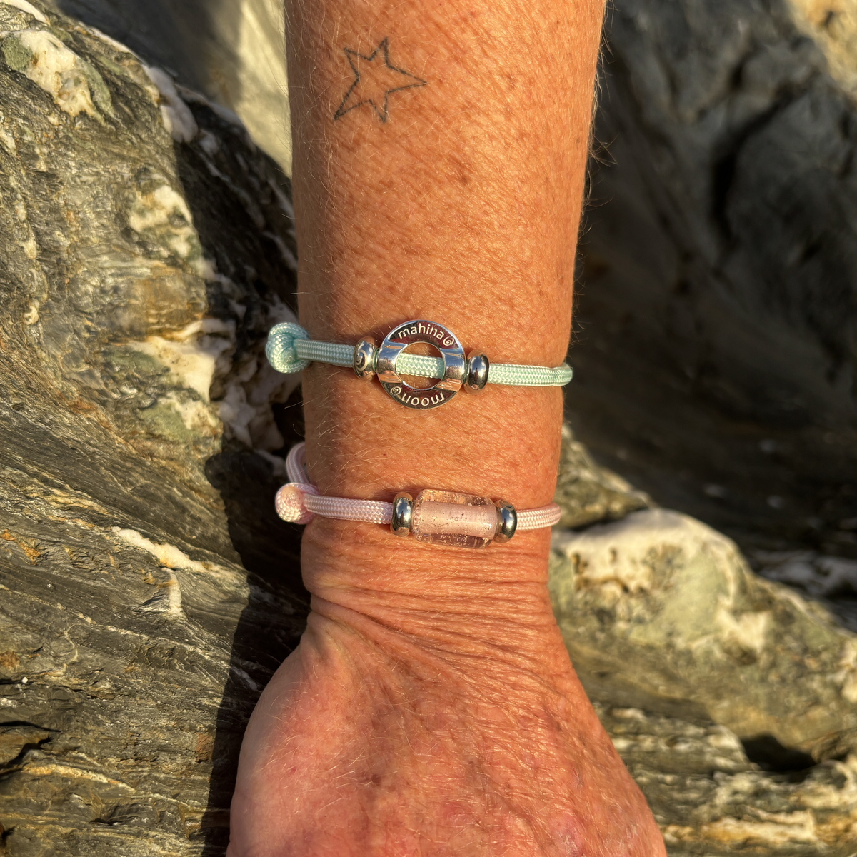 Two delicate beaded bracelets worn on a person's wrist, photographed outdoors against dark rocks. The top bracelet is mint green with a silver-toned round charm engraved 'moon0 and 'mahina'.