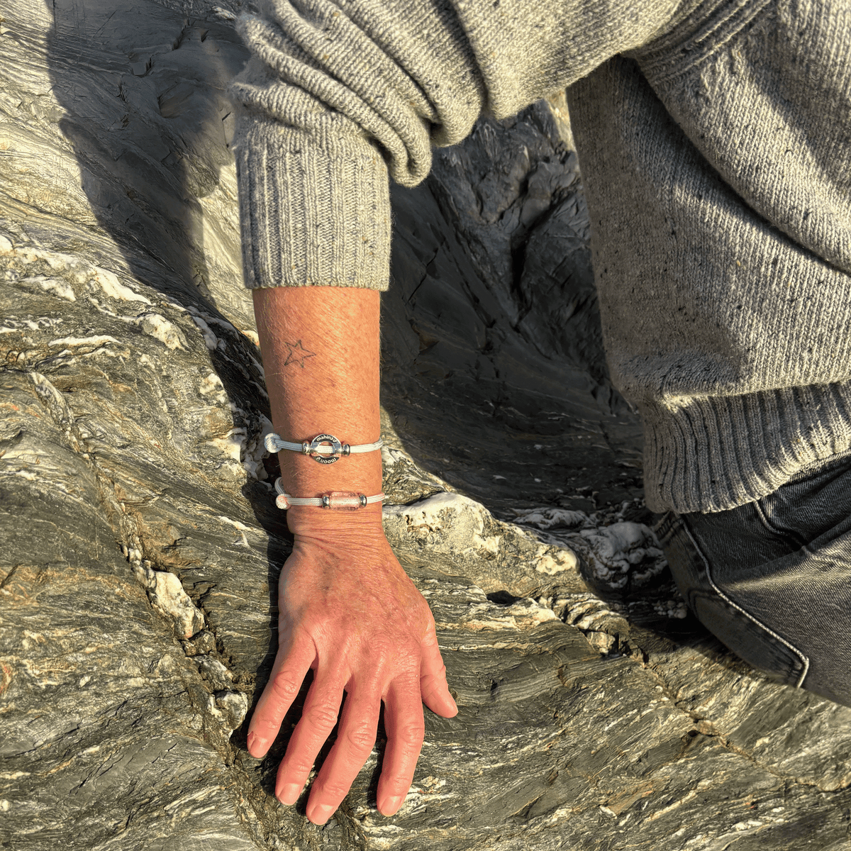A close-up of a person's hand resting on a textured gray rock, wearing two silver-toned bracelets, one of which features a pink stone accent.