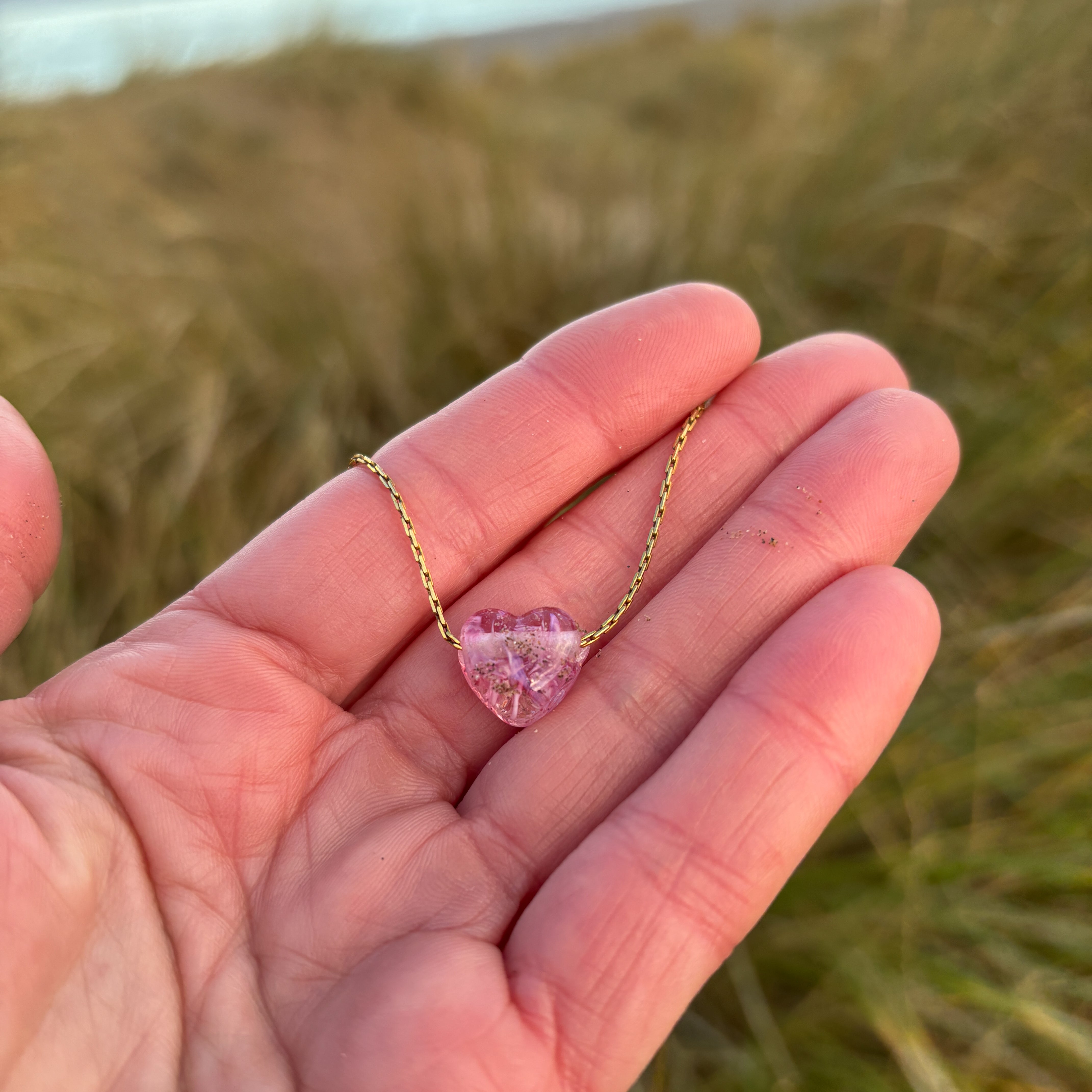 Pink sand heart on gold chain held in persons hand at the beach.