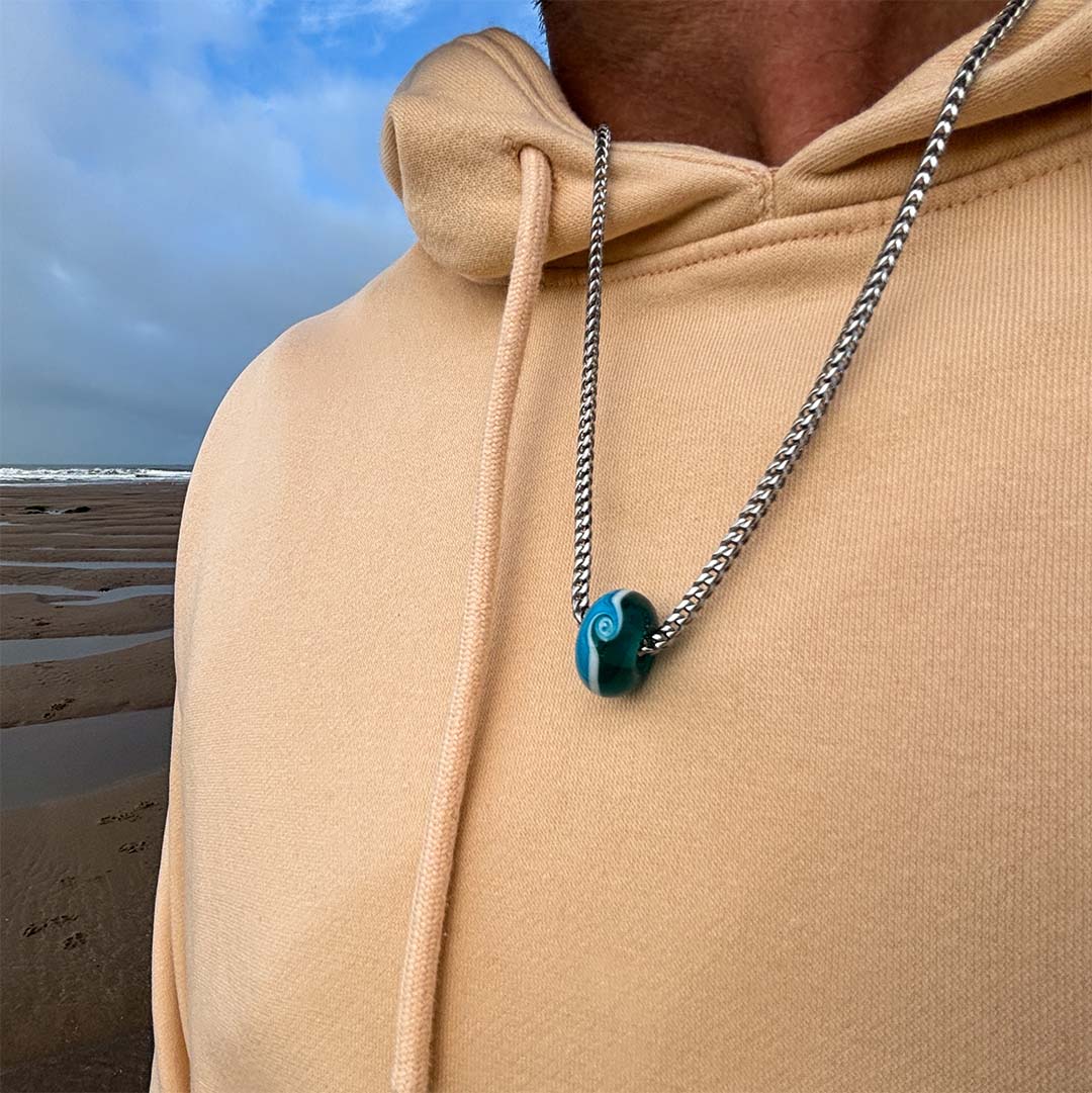 Man on the beach in a apricot coloured sweatshirt wearing a silver necklace with turquoise glass bead.