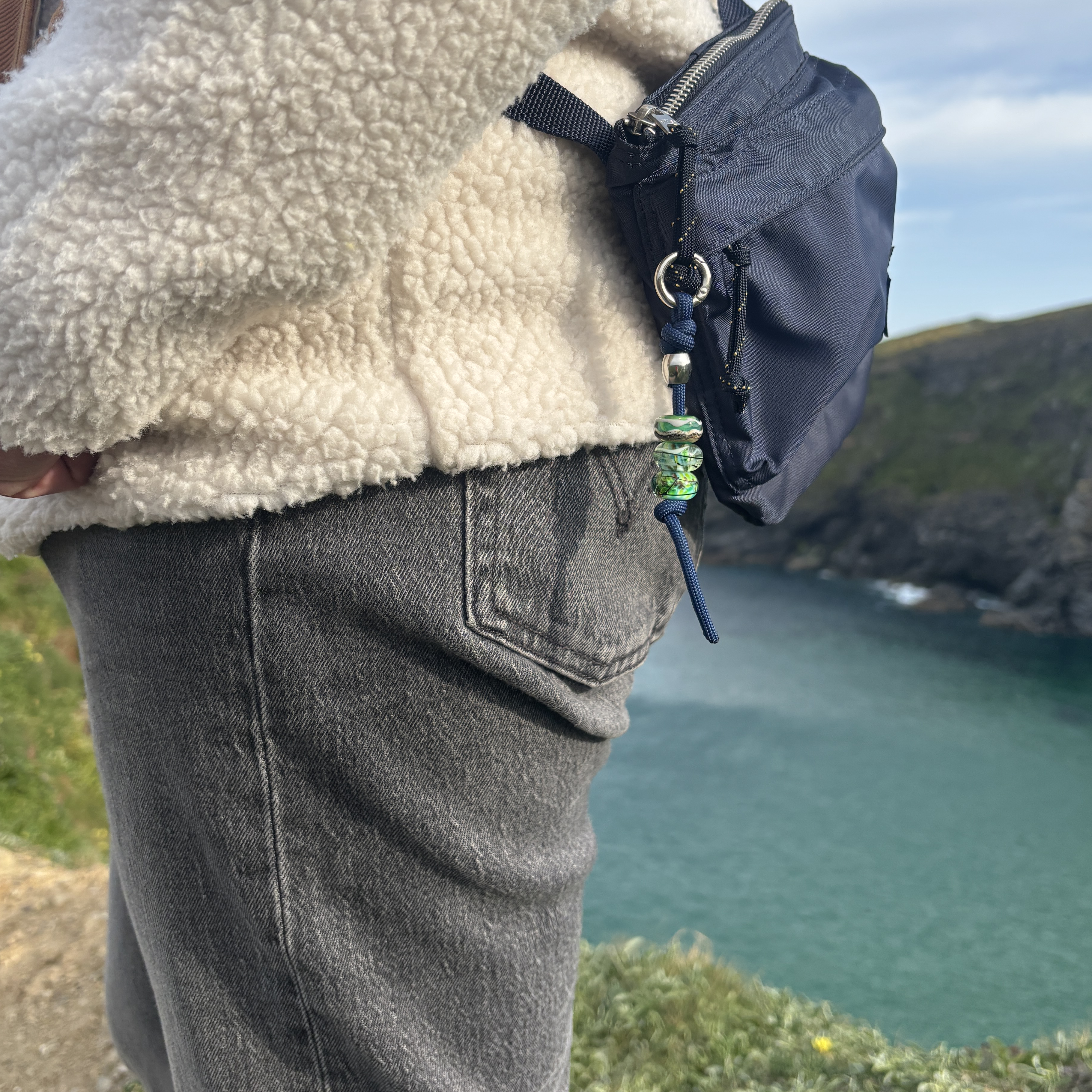 Person wearing a beige fleece jacket with a blue bag over their shoulder, standing on a cliff overlooking the sea.
