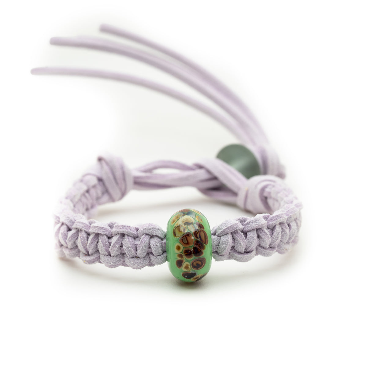 A light purple macrame bracelet with a green glass bead featuring brown and gold spots, set against a plain white background.