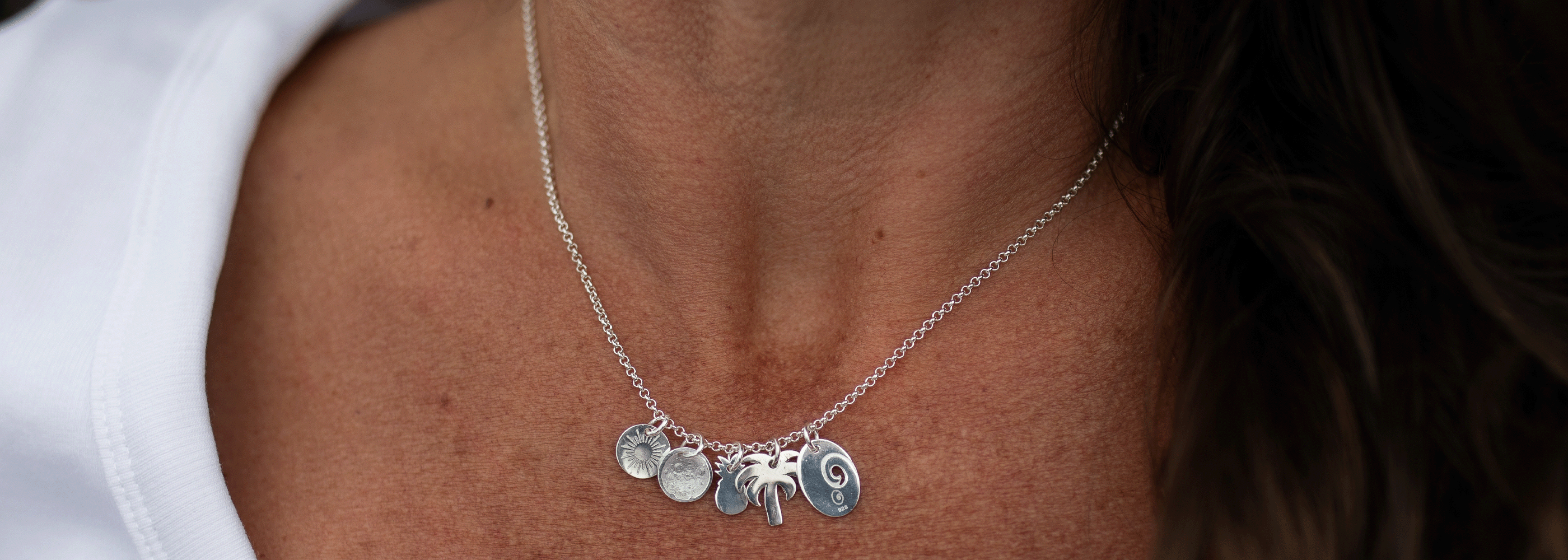 A close-up of a person wearing a delicate silver chain necklace with several small silver charms, including a palm, a crescent moon, and a circular disc, against a white top.