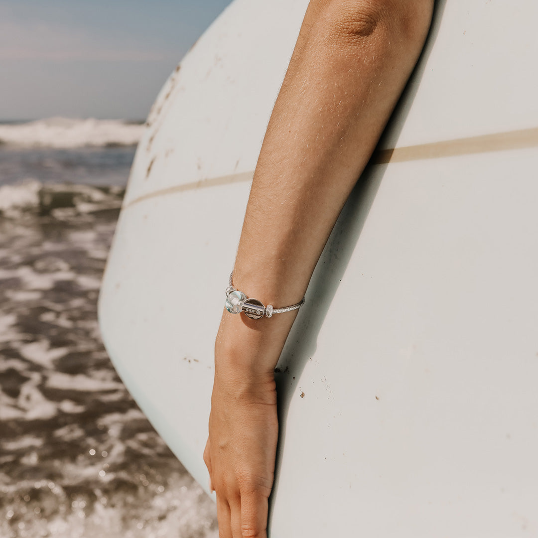 Person carrying a surfboard in to the sea wearing a silver bracelet with glass bead.