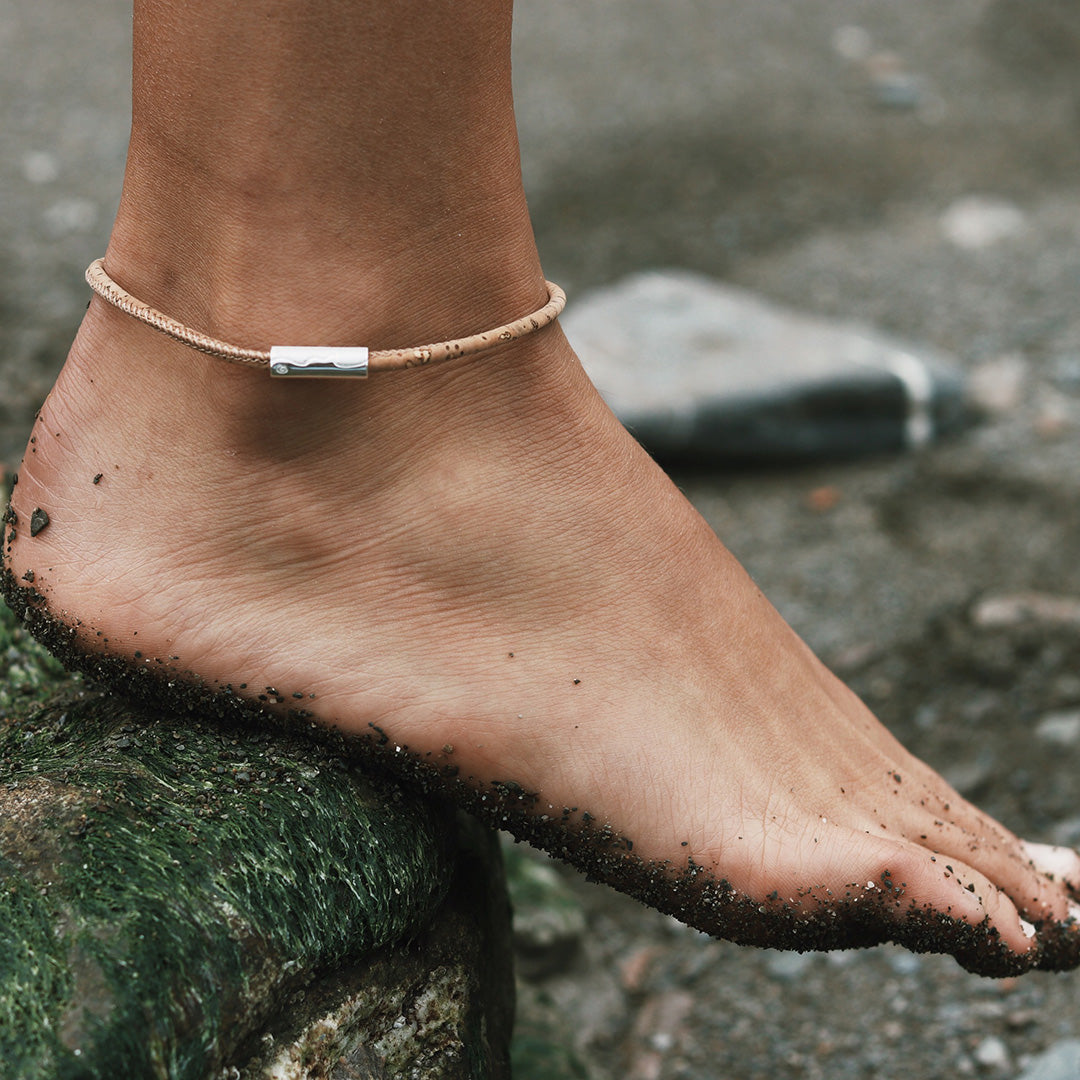 Person wearing a cork anklet with a silver tube charm on it, standing on a rock at the beach.