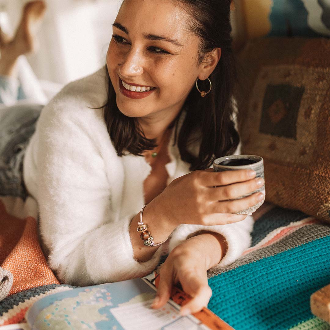 Woman in camper van wearing a silver beaded bracelet, holding a cup of coffee, reading a book.