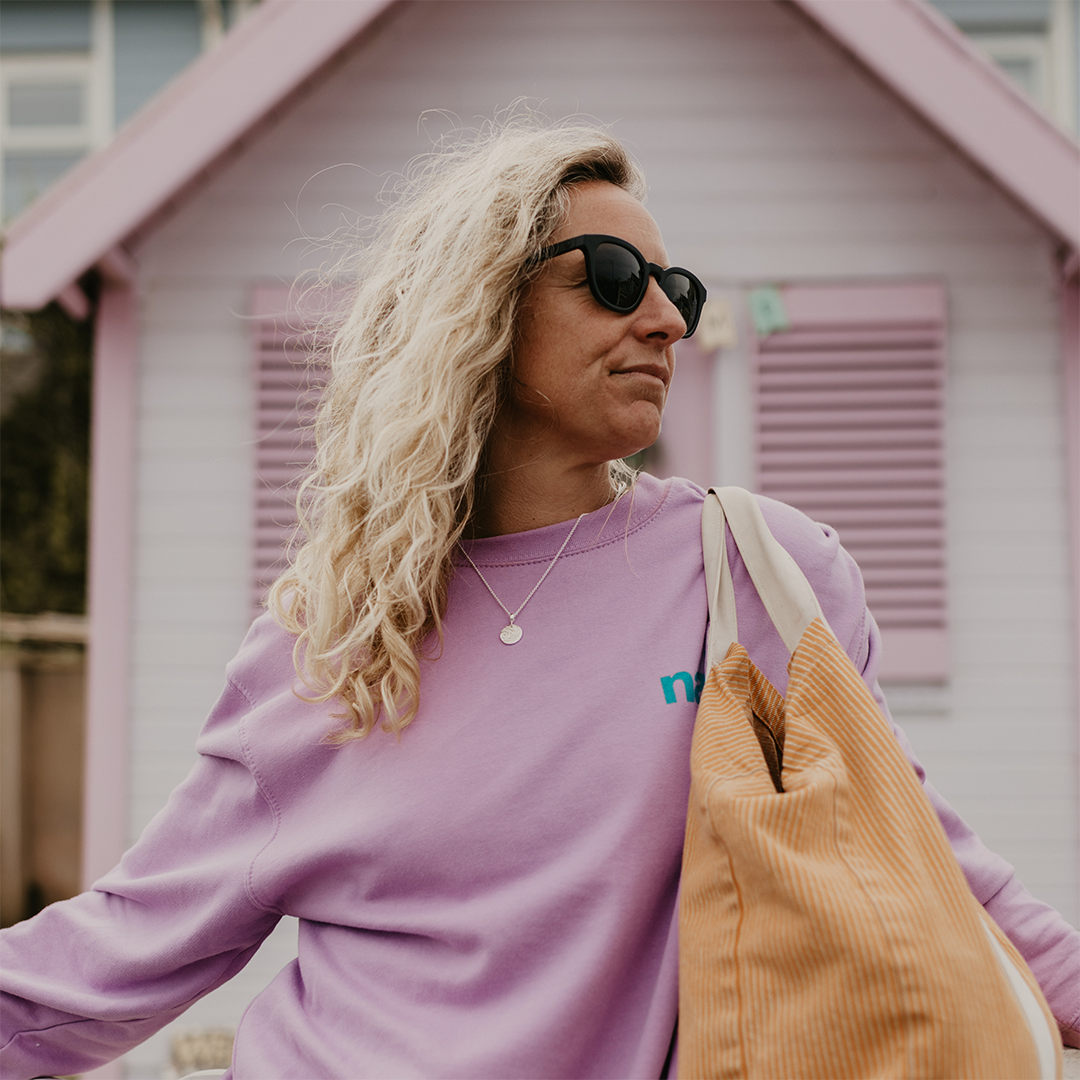 Woman in purple sweatshirt standing in front of a beach hut carrying a brown stripe bag on her shoulder.