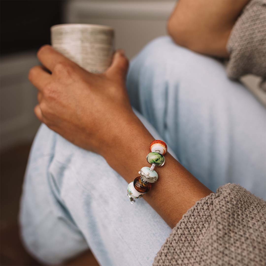 Person holding a coffee cup wearing a brown knitted jumper and wearing a silver beaded bracelet.