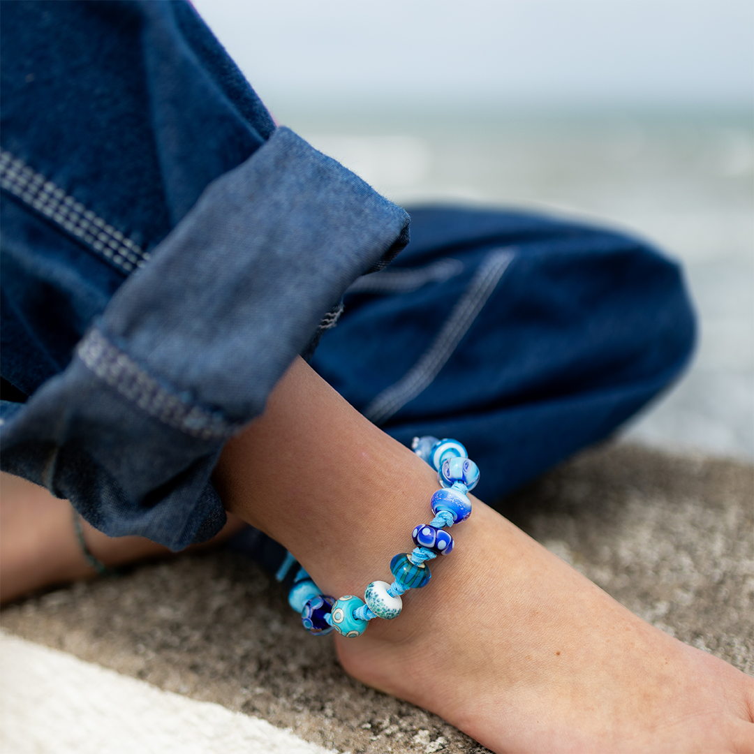 Person sitting cross legged in dark jeans wearing a blue beaded anklet.