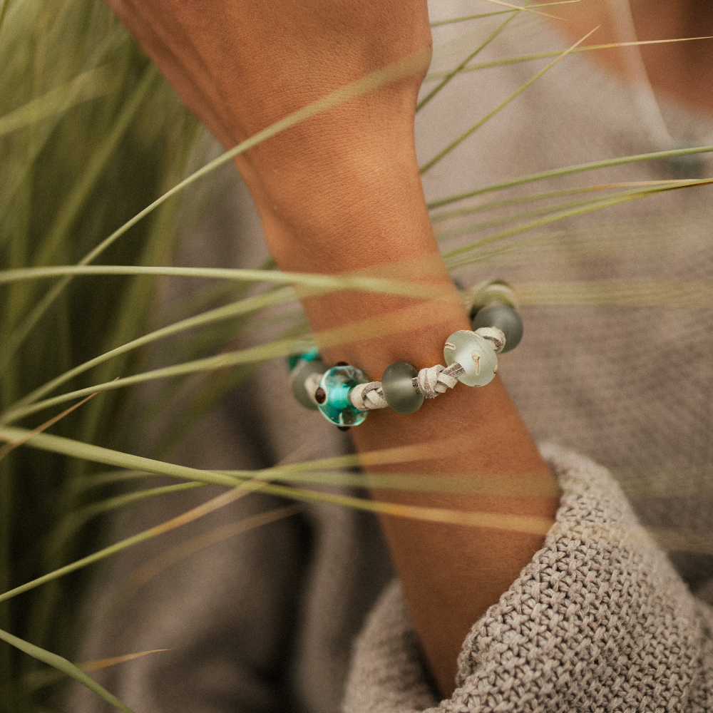 Person wearing knotted grey cord bracelet with patterned Murano glass beads amongst the dune grass.