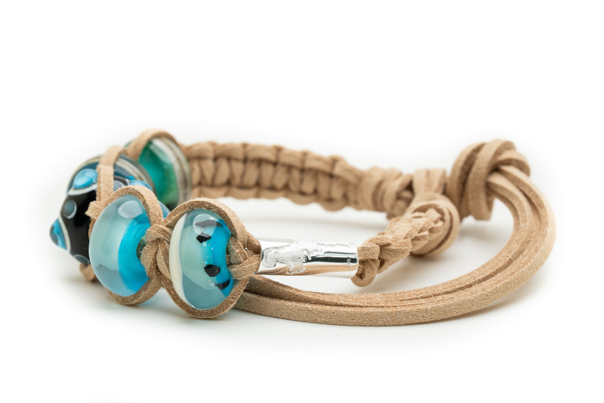 A tan suede wrap bracelet with three decorative glass lampwork beads in shades of blue, black, and green, displayed on a clean white background.