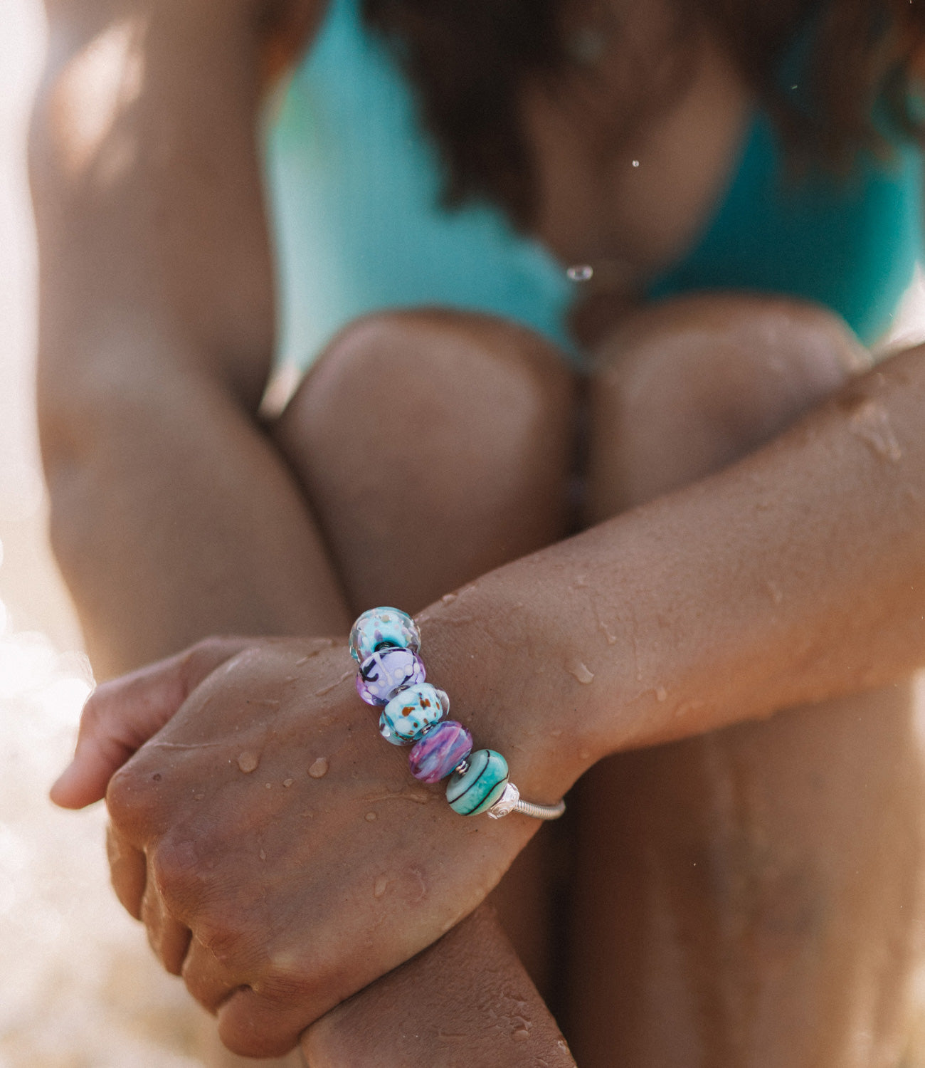 Green, blue and purple glass beads worn on silver snake chain bracelet by surfer girl at beach in teal bikini.