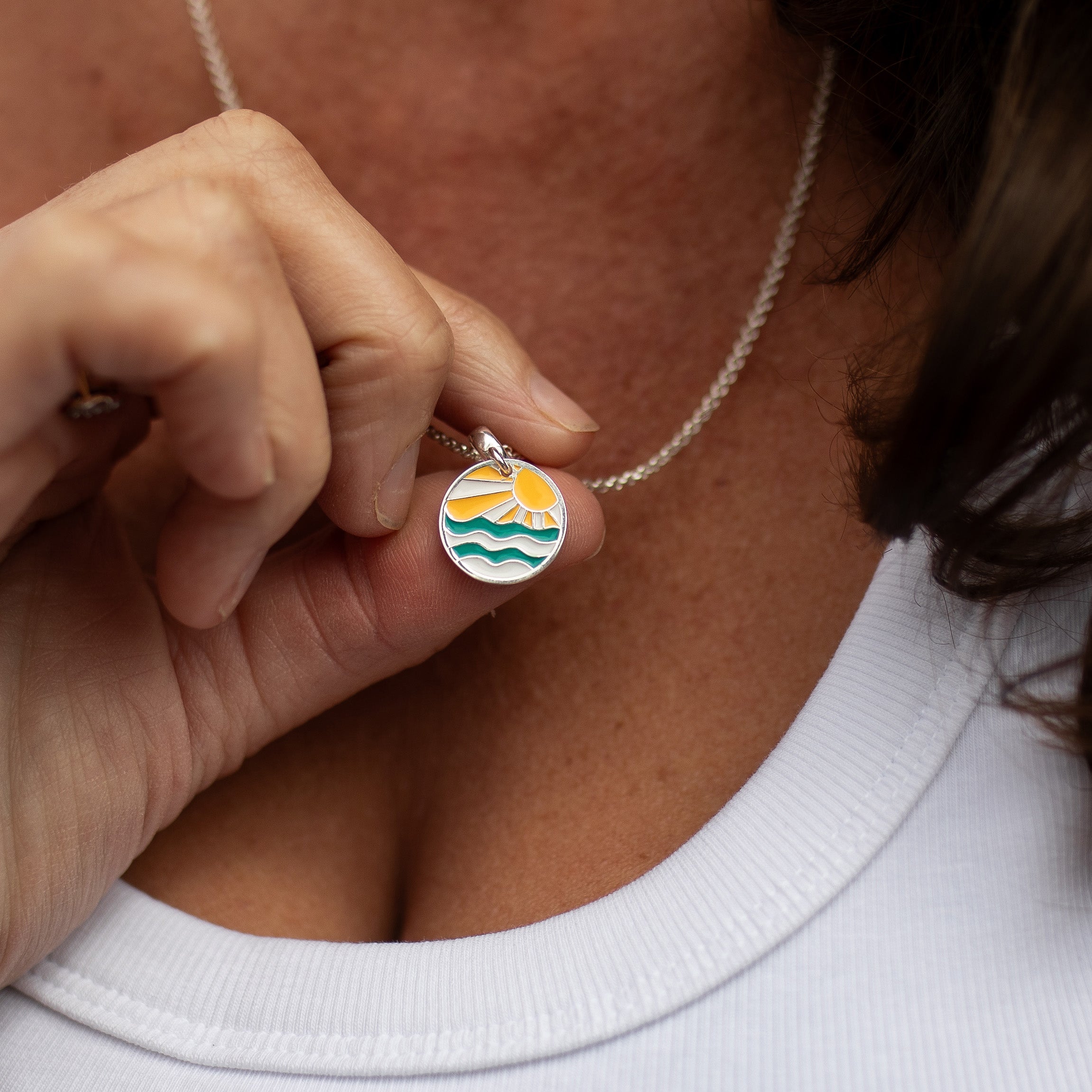 A close-up lifestyle shot of a person holding a small round silver pendant necklace. The pendant features a colorful enamel design of a yellow sun over green and blue waves.