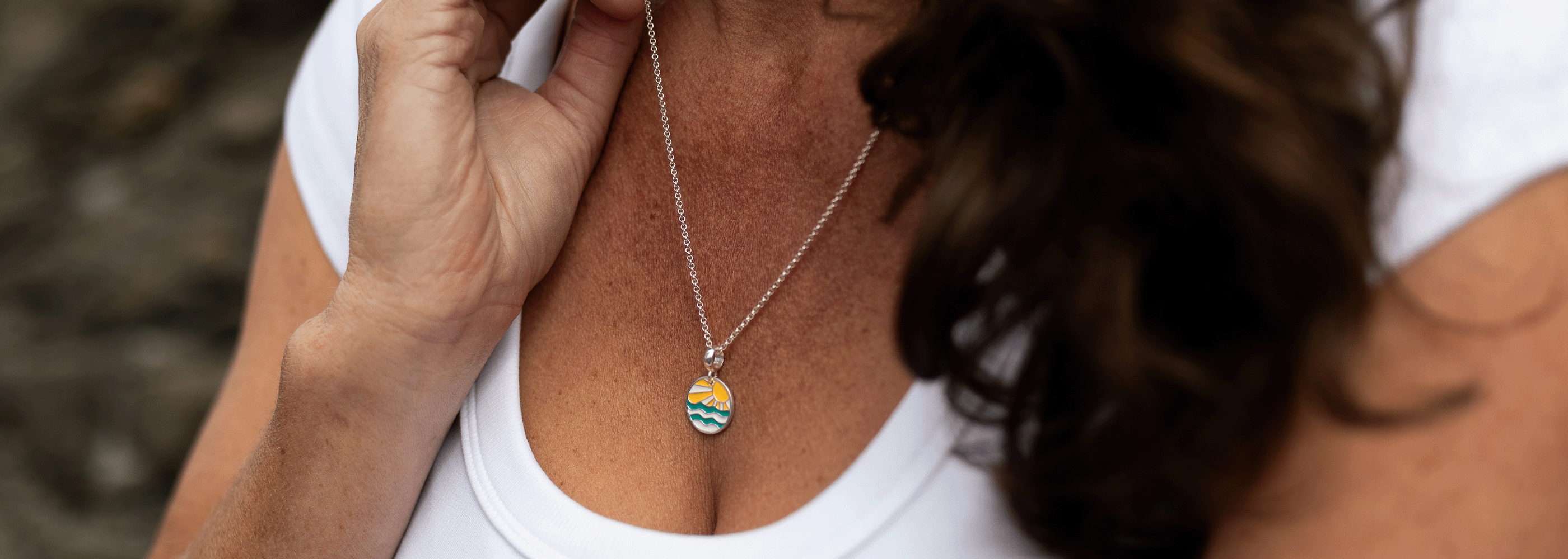A close-up lifestyle shot of a woman wearing a silver chain necklace with a small oval pendant featuring a colorful landscape design.