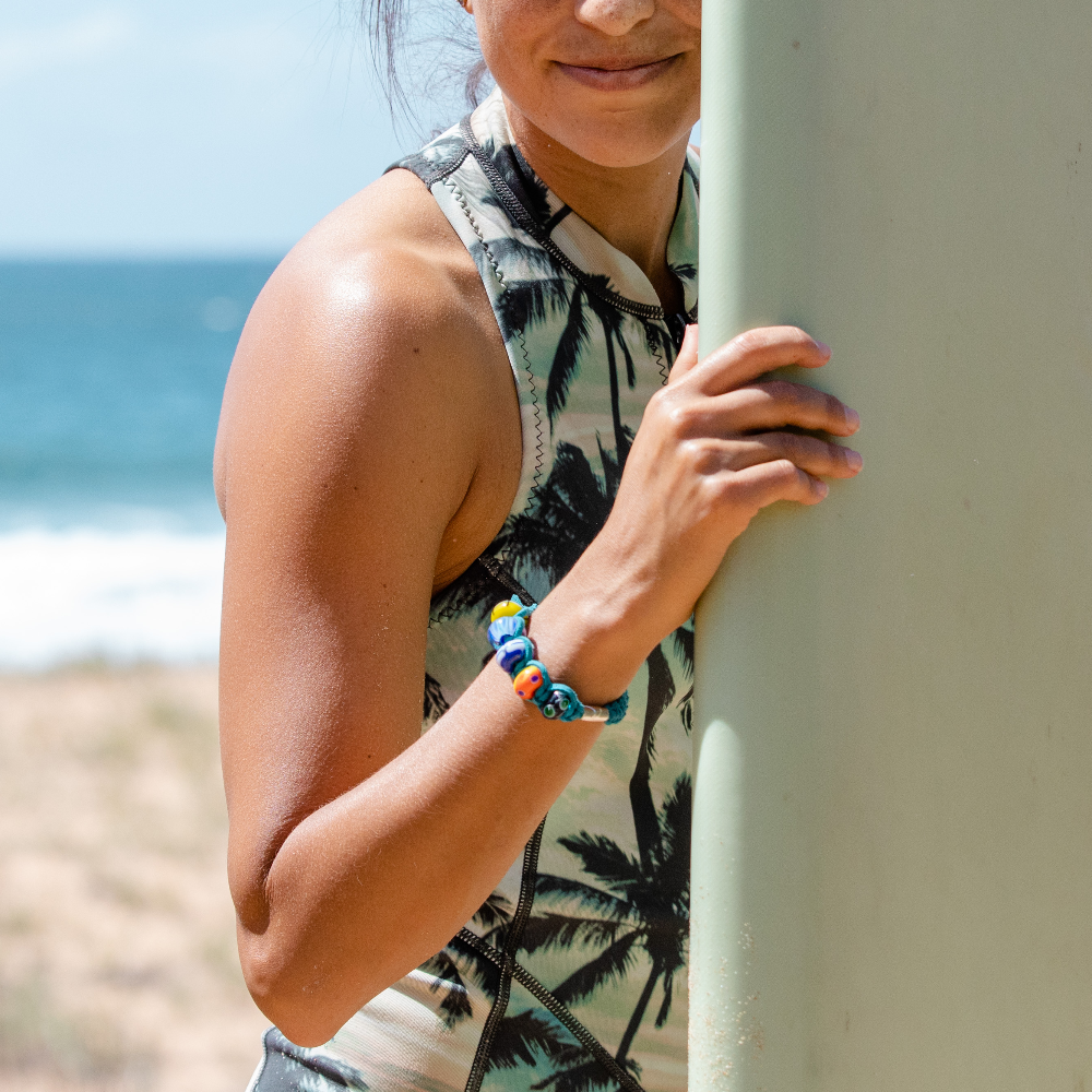 Woman surfer holding her surfboard in front of her at the beach wearing a palm print swimsuit and a teal cord beaded bracelet.