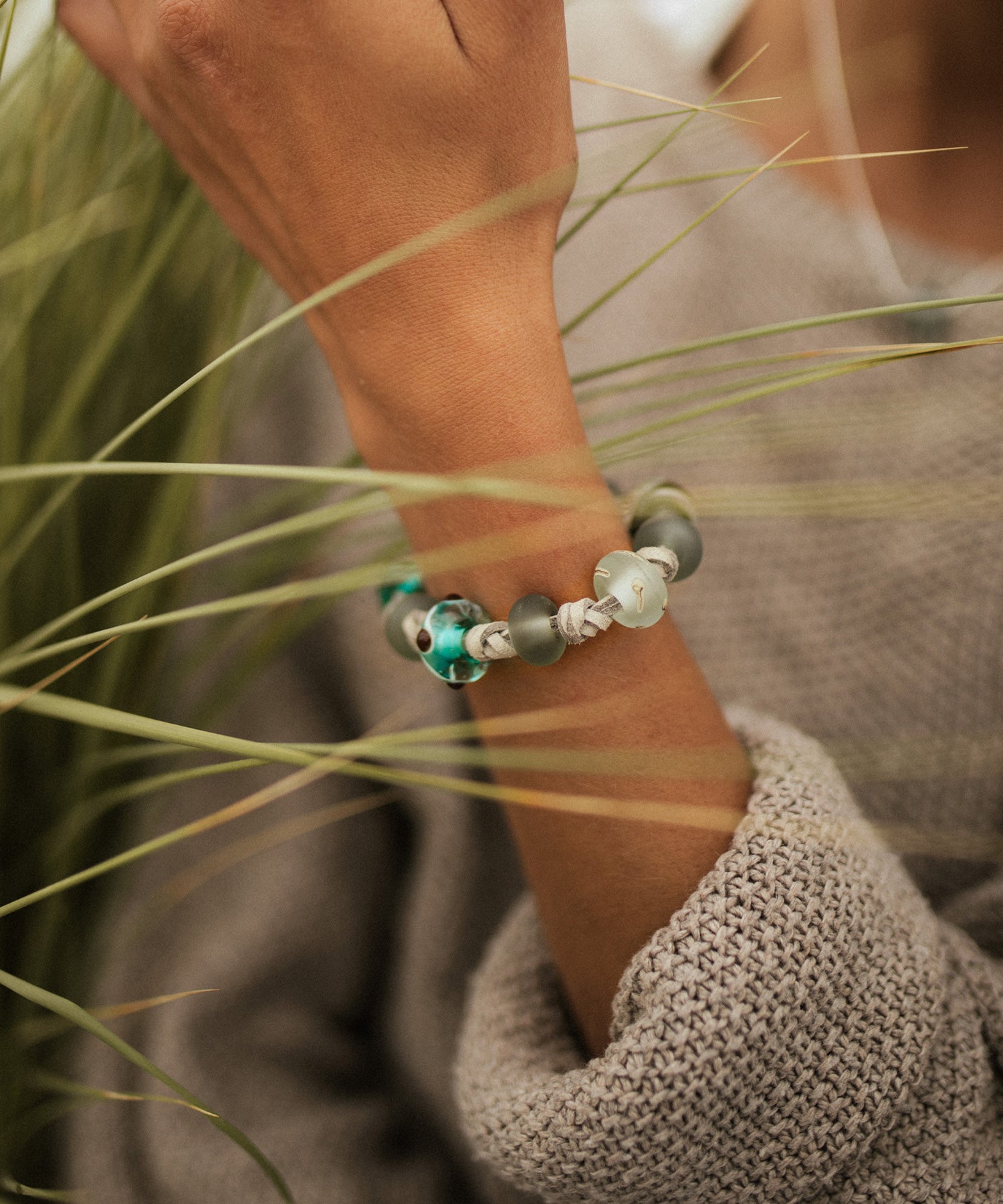 Devon surf beads on grey cord bracelet worn by woman wearing brown knitted jumper on the beach.