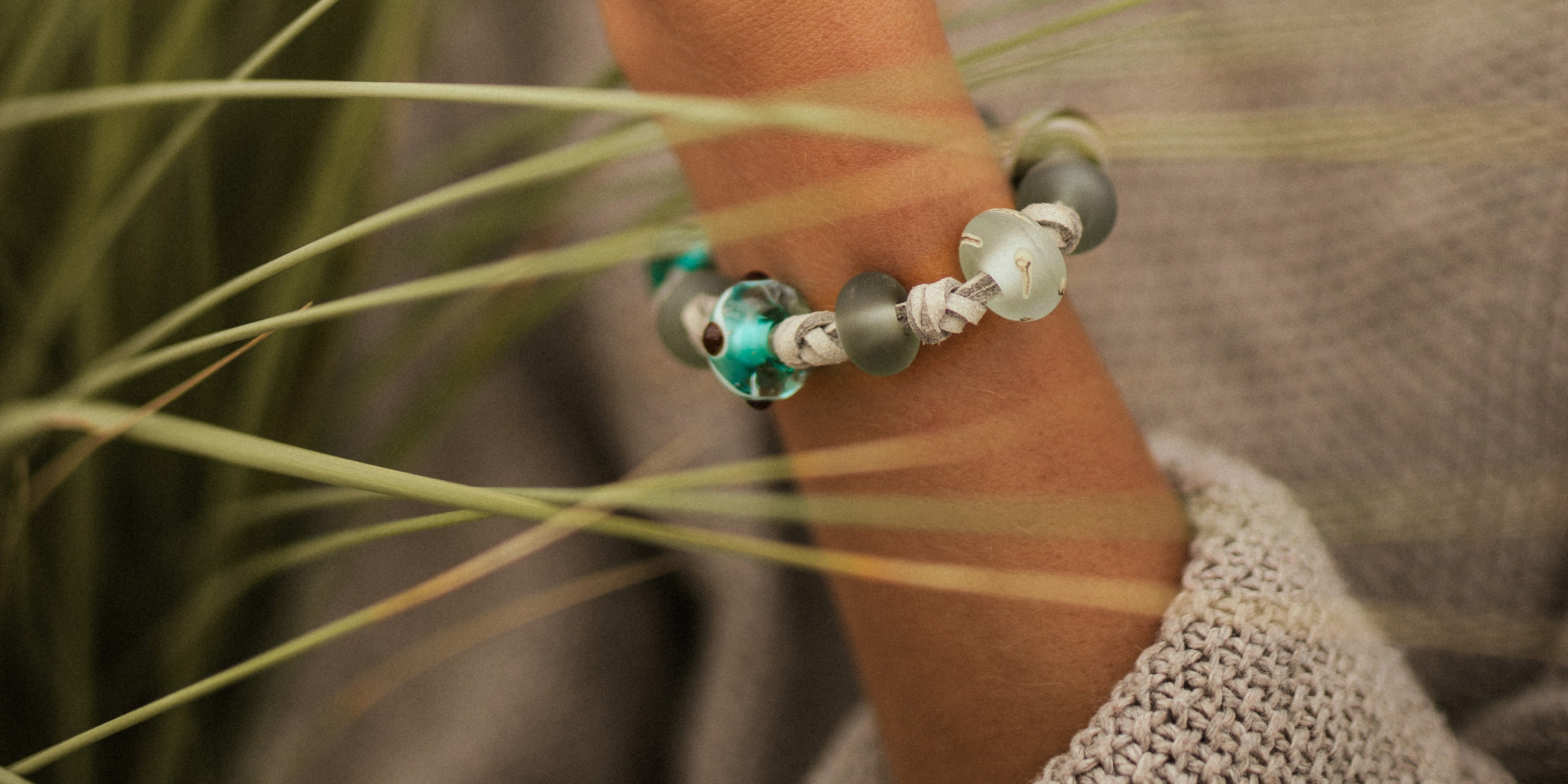Devon surf beads on grey cord bracelet worn by woman wearing brown knitted jumper on the beach.