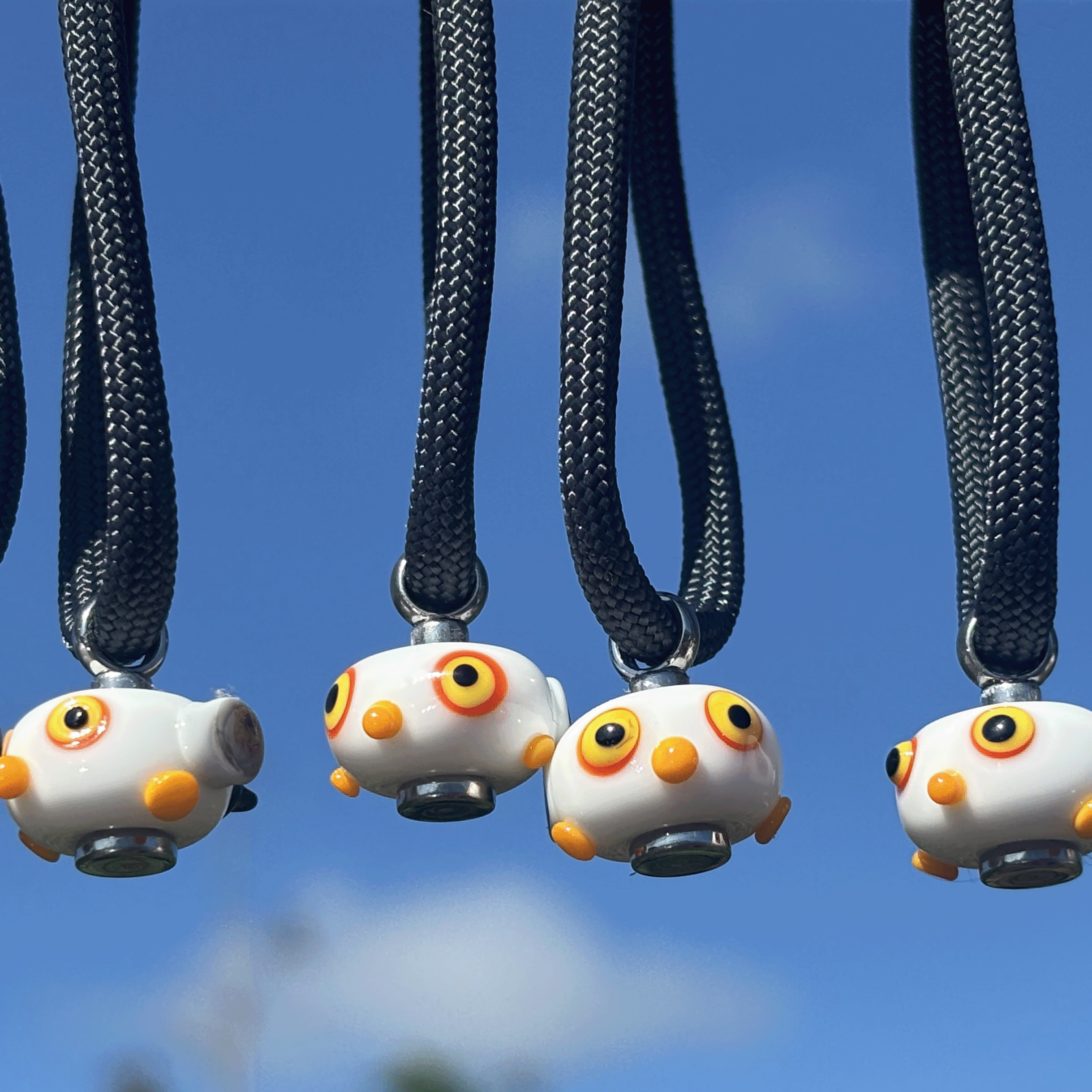 Four small white seagull-shaped pendant necklaces with black braided cords, hanging against a bright blue sky background.